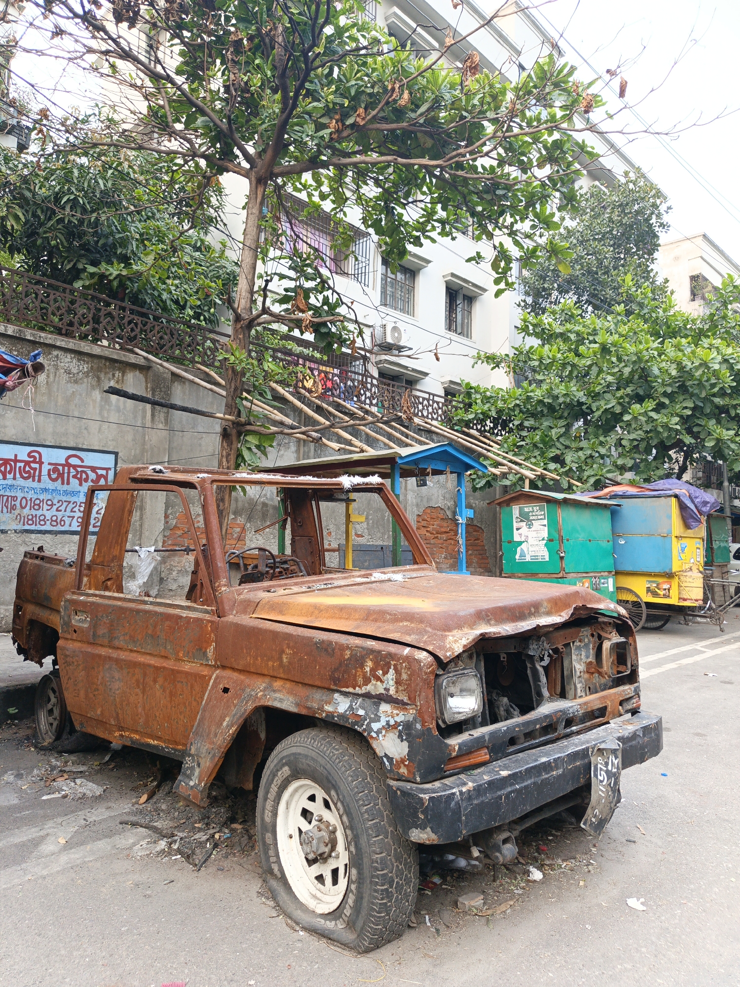 The image shows a heavily rusted and dilapidated pickup truck parked on a street in Dhaka, Bangladesh. The truck is missing most of its body panels, windows, and interior components, and it is covered in a thick layer of rust. Its tires are flat. The surroundings include a building with a tree in front, some street vendors' stalls, and a sign in Bengali that reads "Kazi Office". The stalls, a brick wall with fencing on top, and more trees line the background. The general environment suggests an urban setting with a mix of commercial and residential elements.