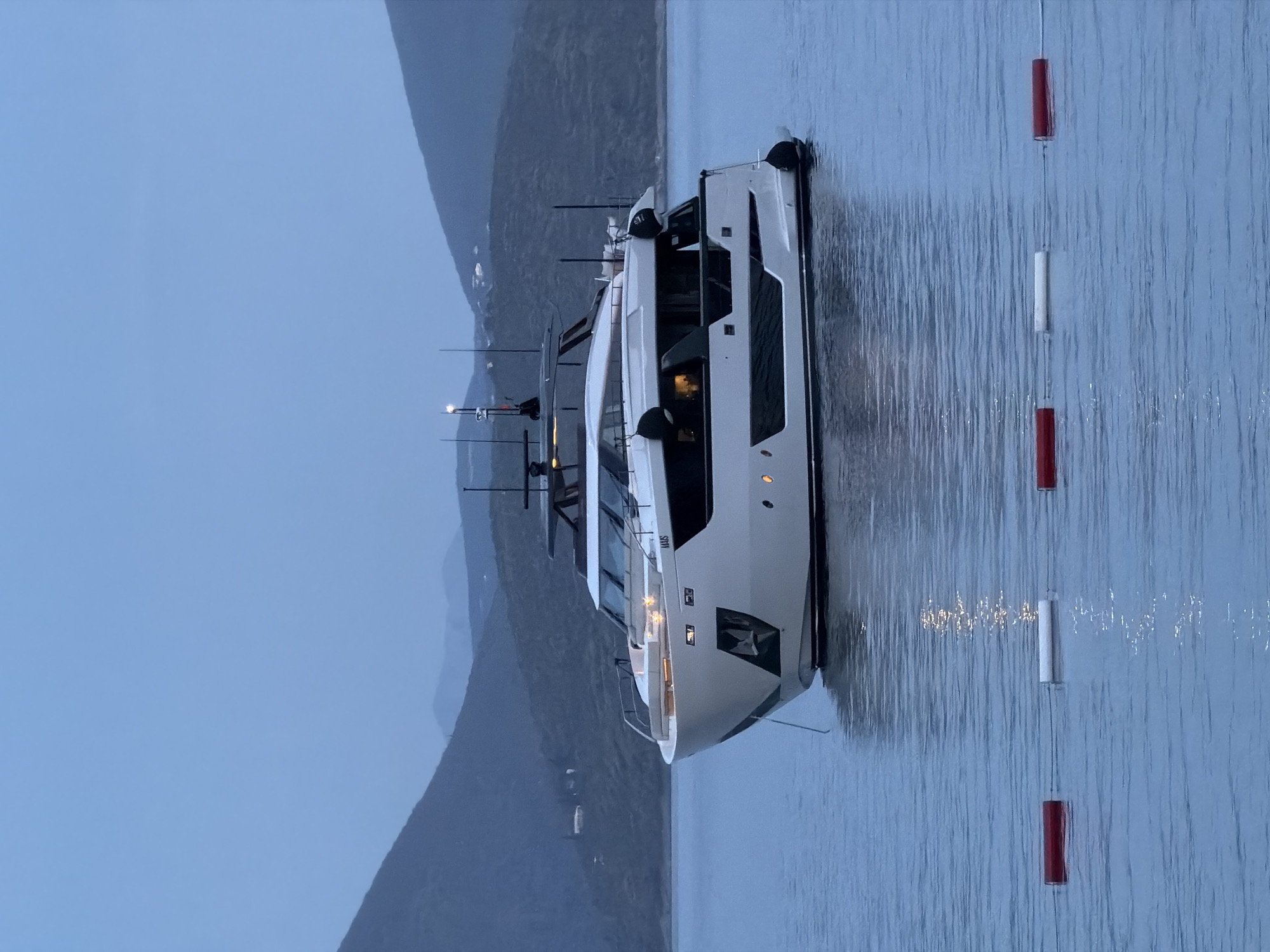 A large white yacht is docked in the waters off Lješevići, Montenegro. The vessel is oriented lengthwise in the frame, with its bow pointing towards the lower right corner. Mountains can be seen in the background. A series of red and white buoys mark off the area in the water to the right of the yacht. The sky is overcast.