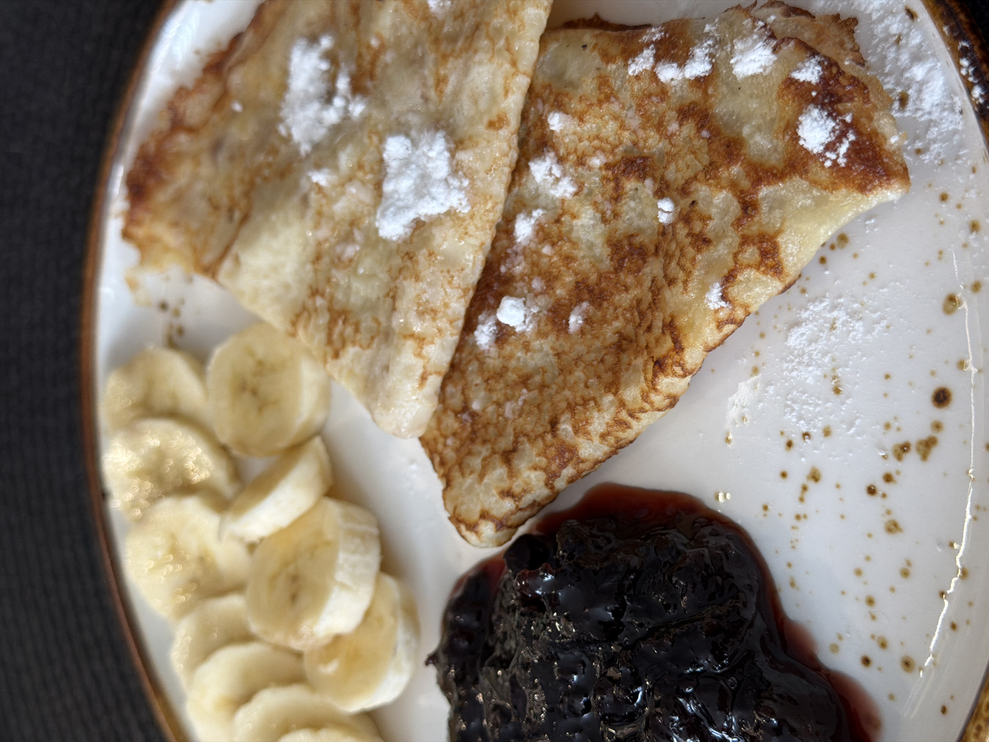 A top-down shot shows a plate of food, likely a dessert. Two crepes folded into triangles are dusted with powdered sugar. Sliced bananas are arranged in a curve along the left side of the plate. A dark, glossy jam or compote is positioned near the bottom right. The plate has a speckled pattern and a brown rim. The surface beneath the plate is dark and textured. The scene is presumed to be in Засечное, Russia.