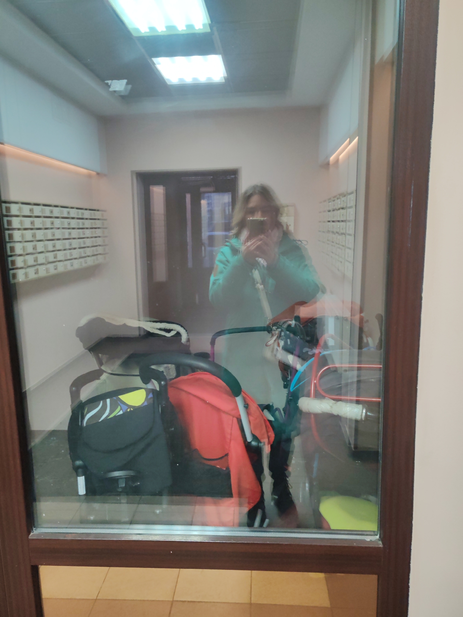 The image captures a reflection of a person taking a selfie in what appears to be an indoor hallway or an apartment building entrance in Saint Petersburg, Russia. The shot is taken through a large, rectangular glass panel set in a dark brown wooden frame, which reflects the person and objects in the foreground, while also allowing a view of the background.
The main subject is a person, likely female, with medium-length hair, wearing a teal or green hooded jacket. They are holding a smartphone to take a selfie, with their face obscured by the device. To the left and in front of the person is a baby stroller, primarily black with a colorful pattern (yellow, blue, green) on its fabric, and some red fabric components. To the right of the stroller, partially visible, is another item of children's transport, possibly a scooter or small bicycle, with white handlebars.
The setting is an indoor corridor. In the background, visible through the glass, there are multiple rows of white, wall-mounted mailboxes on the left wall. A dark-framed doorway is seen in the center background. The ceiling features bright fluorescent light panels, illuminating the space. A subtle horizontal light fixture or decorative trim runs along the upper part of the left wall. The floor is covered with light-colored tiles. No discernible text is visible in the image.