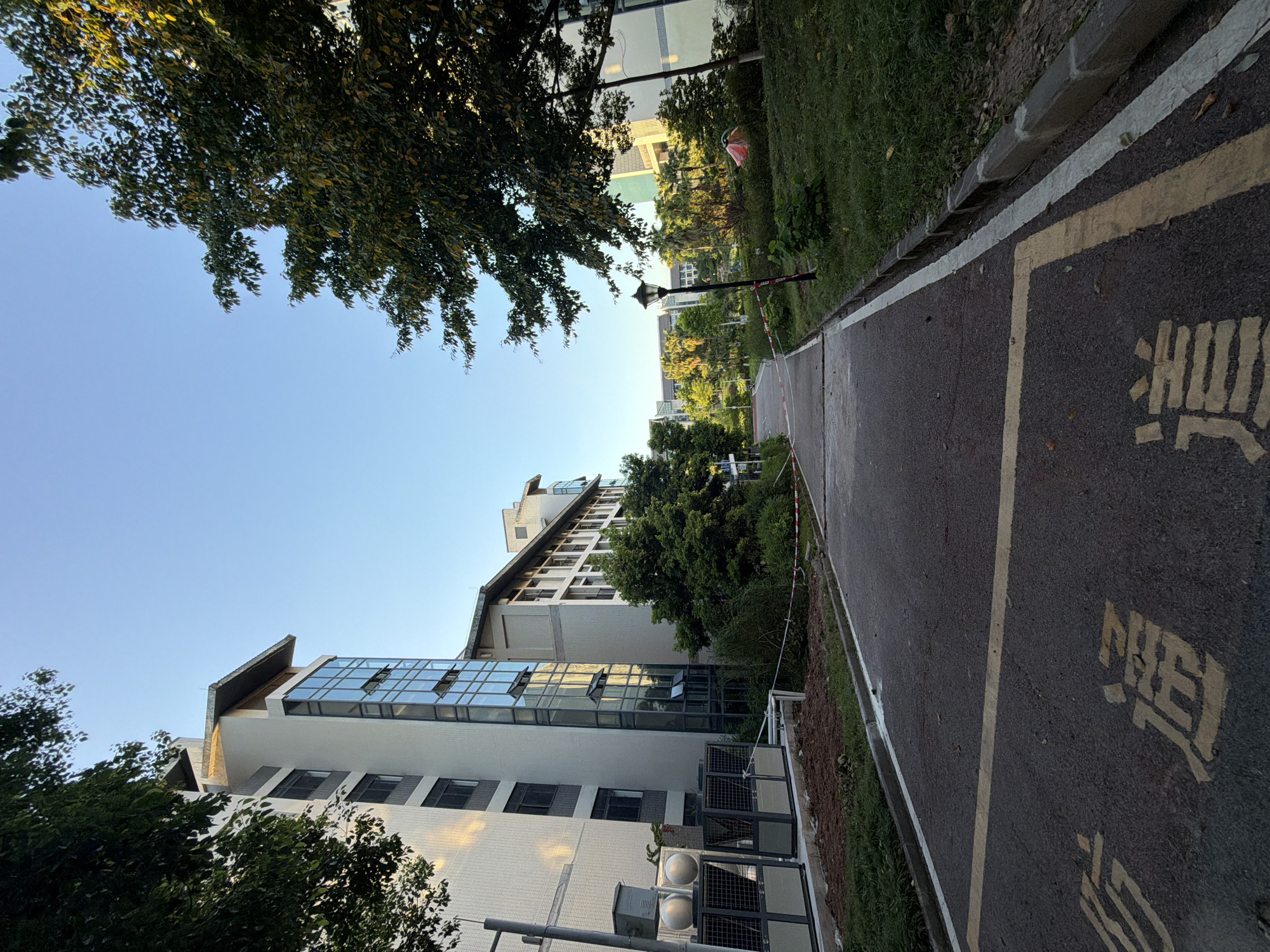 The image depicts an outdoor scene in Jiangjin, China, likely on a university campus or a similar institutional setting. A paved pathway, marked with yellow lines and Chinese characters, leads into the distance. On the left, a modern, multi-story building with white brickwork and large glass windows stands prominently. Adjacent to the building are various mechanical units and some unkempt earth. To the right of the pathway, a grassy area is interspersed with trees and a lamppost. Further down the path, more buildings and lush greenery are visible under a clear blue sky. Red and white caution tape is strung across the path in the middle ground, suggesting a restricted area. The overall atmosphere is serene, captured during daylight hours.
