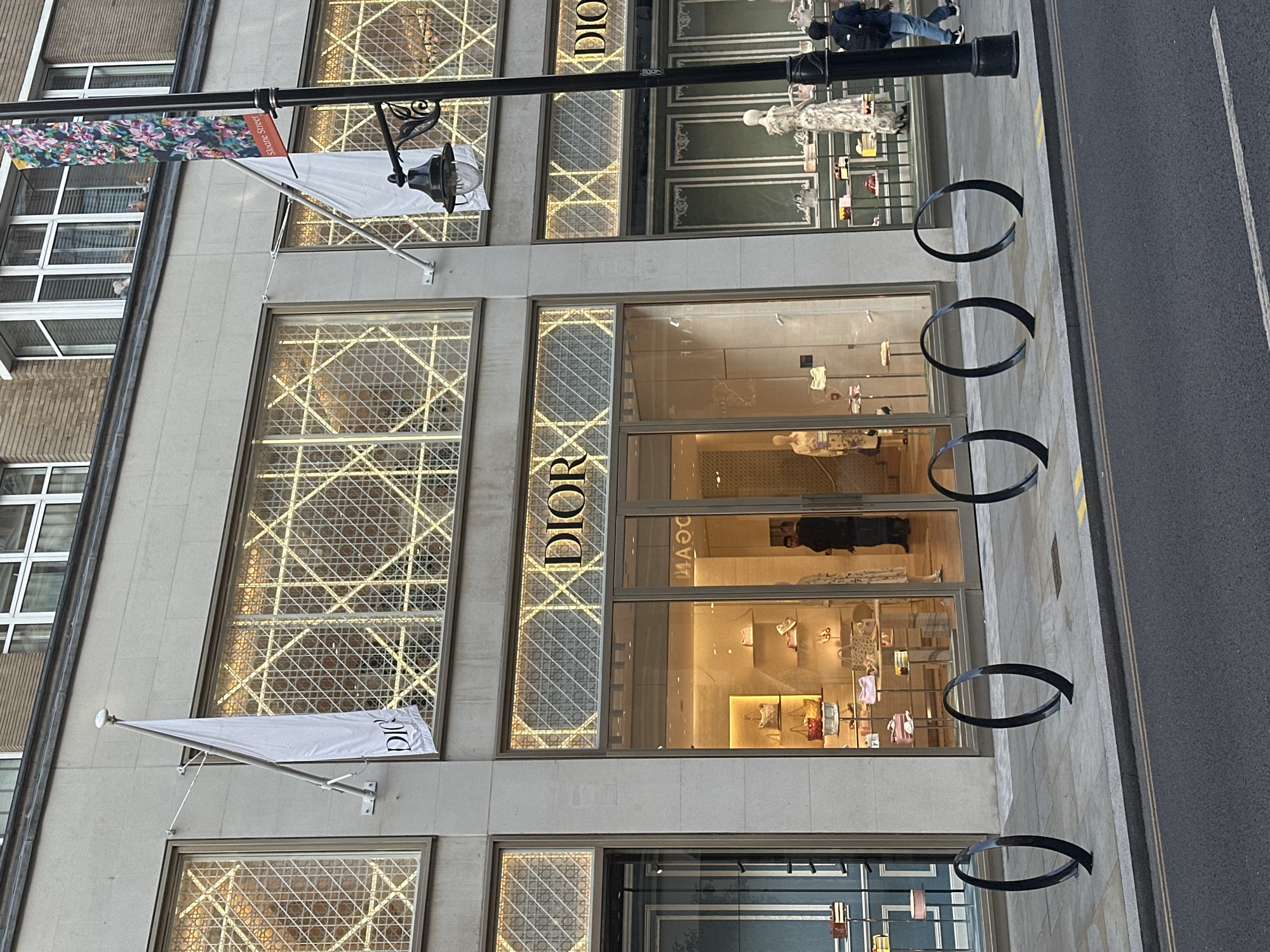 The image shows the facade of a Dior store on a city street. The store has large display windows showcasing handbags and mannequins dressed in clothing. The Dior logo is prominently displayed above the entrance. A white flag with the Dior logo hangs from a flagpole on the left side of the building. A lamppost with a hanging light fixture stands in front of the store. The street in front of the store has a sidewalk with black, circular bicycle racks. A few people are visible walking past the store. The building appears to be made of stone and has large windows on upper floors. The overall scene suggests a high-end retail environment in an urban setting.