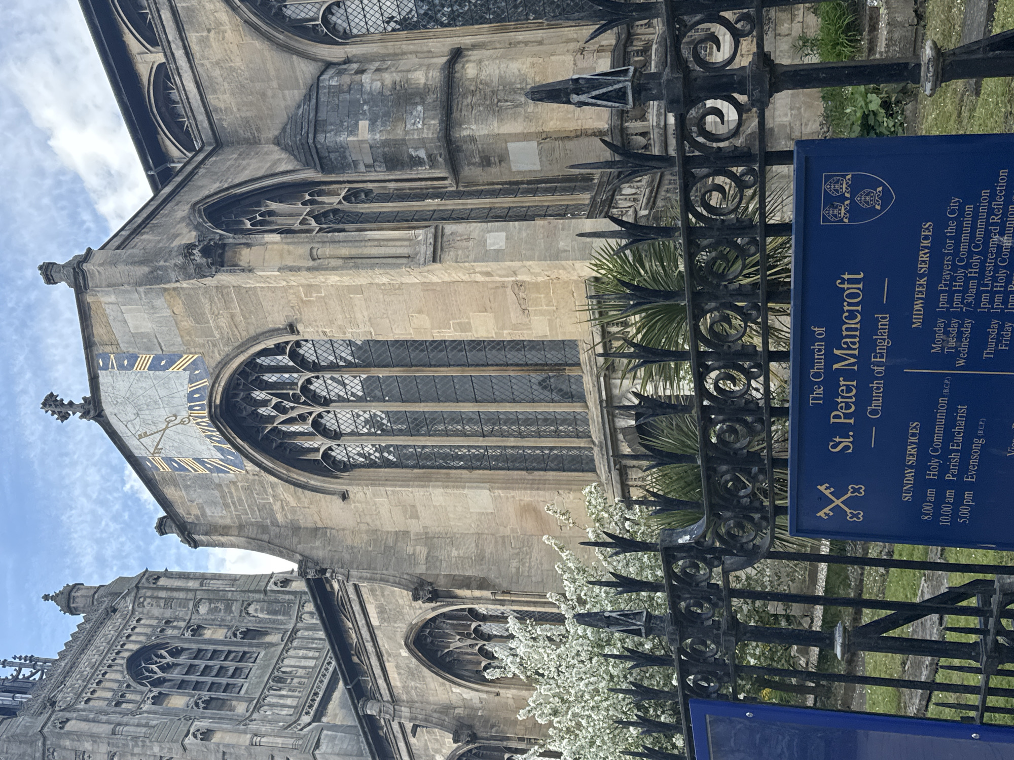 The image captures the Church of St. Peter Mancroft in Norwich, United Kingdom. The photograph is taken from outside the church, looking up at the building's facade. A wrought-iron fence runs across the foreground. Behind the fence is a sign detailing the church's service times. The church architecture is visible, featuring large stained-glass windows, a sundial on the wall, and a tower. The sky is partially visible in the background.