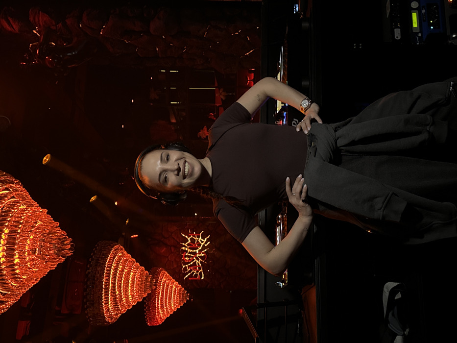 A female subject, appearing to be a disc jockey, stands in a confident pose with hands on hips, smiling, and wearing black over-ear headphones. She wears a dark maroon t-shirt, with a dark grey sweatshirt tied around her waist, and dark grey pants. A gold-toned watch is visible on her right wrist, and rings are on her left hand. She stands in front of a DJ booth equipped with professional audio mixers and turntables. The scene is illuminated by red and orange ambient lighting. Three multi-tiered crystal chandeliers glow with a reddish-orange hue, suspended from the dark ceiling in the upper left. Spotlights emit red beams. In the mid-background, a neon sign displaying a stylized word, possibly "ЛЁД," glows orange-red on a dark, textured wall. To the right, a rack of audio equipment with illuminated blue and green indicators is visible. This interior scene depicts a nightclub or lounge environment in Moscow, Russia.