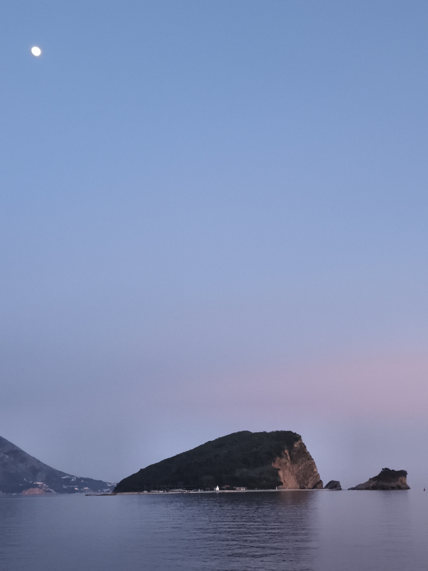 The image shows a twilight scene in Budva, Montenegro. The sky is a gradient of blue to pink. In the upper left is the moon. The foreground features a body of water with islands. The largest island is covered in trees, and a sandy beach with buildings is visible. In the distance to the left of the largest island is a coastline with structures. To the right of the island are smaller rocky islands. The sea reflects the colors of the sky.