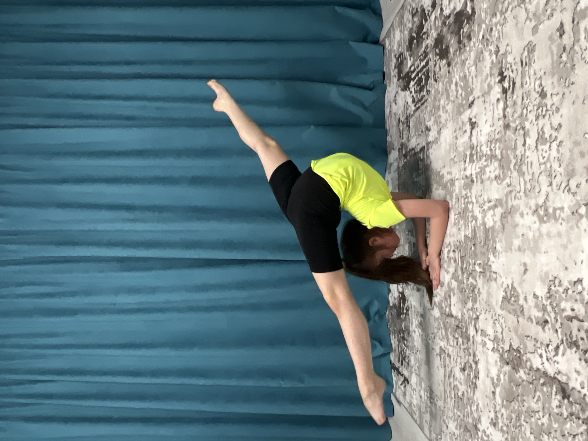A young girl is performing a handstand split against a wall. She is wearing a bright yellow t-shirt and black shorts. Her left leg is raised upwards, and her right leg is extended straight behind her. The wall behind her is decorated with a grey and white patterned wallpaper. To the left of the girl is a teal curtain. The location is unknown. There is no evidence it's located in the city of Chaikovsky, Russia.