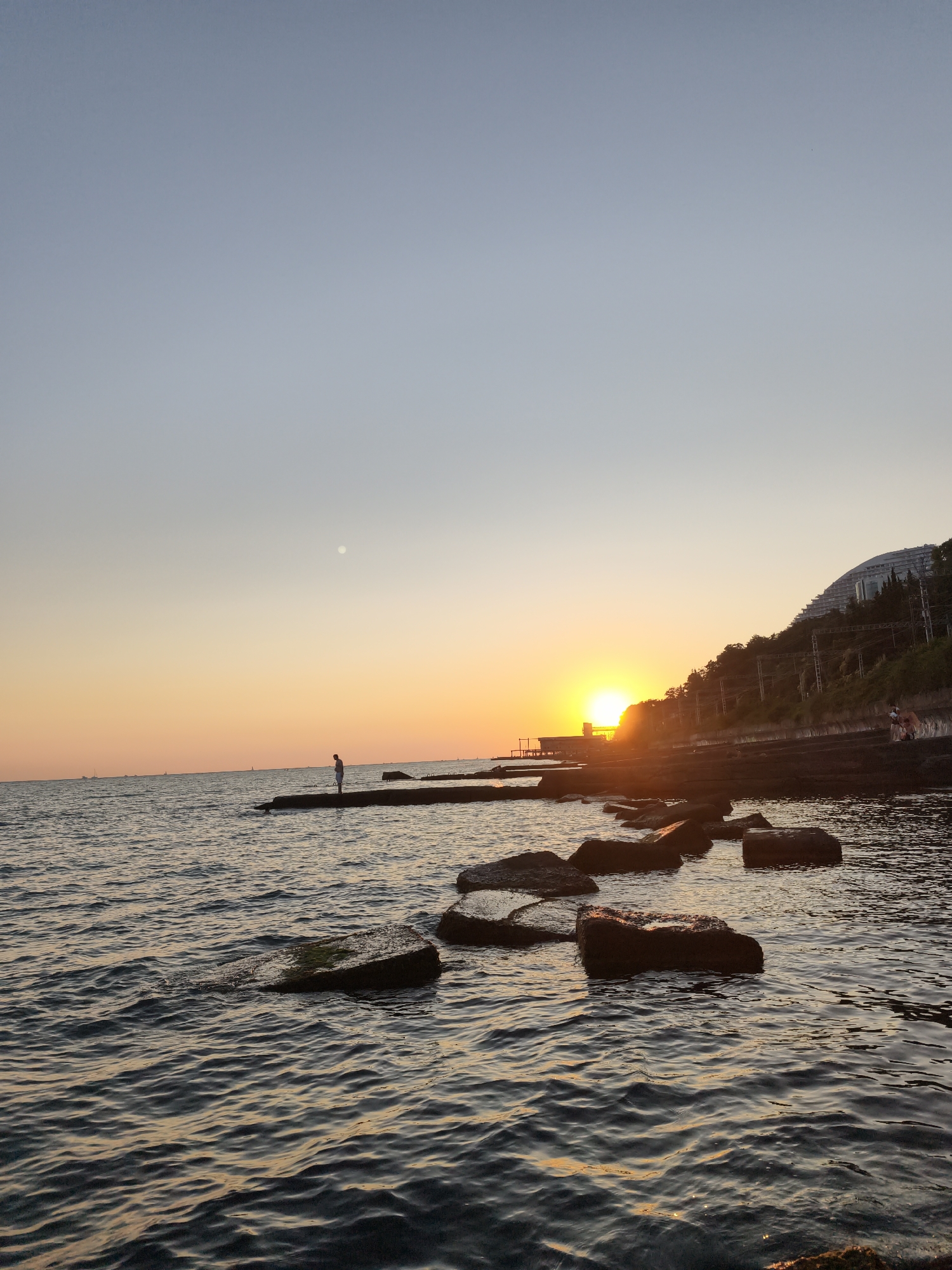 This is a photograph taken at sunset on the coast of Sochi, Russia. The scene depicts a body of water, likely the Black Sea, with a shoreline composed of rocks and concrete structures. The sun is setting on the horizon, casting a warm orange and yellow glow across the sky and water.
In the foreground, several large rocks are partially submerged in the water, their surfaces wet and reflective. The water itself is rippled, with gentle waves lapping against the shore. A concrete pier extends into the water, and a lone individual stands on it, silhouetted against the bright sky. Further along the coastline, more concrete structures form a walkway or breakwater.
To the right of the frame, a steep, tree-covered hillside rises from the coast. Buildings, including a distinctive curved structure, are visible nestled among the trees and on the hillside, illuminated by the setting sun. Overhead power lines and poles are also present along the slope. In the distance, several sailboats can be seen on the water, adding to the tranquil atmosphere of the scene. The moon is faintly visible in the upper left portion of the sky. The overall impression is one of a peaceful evening by the sea in a coastal resort town.