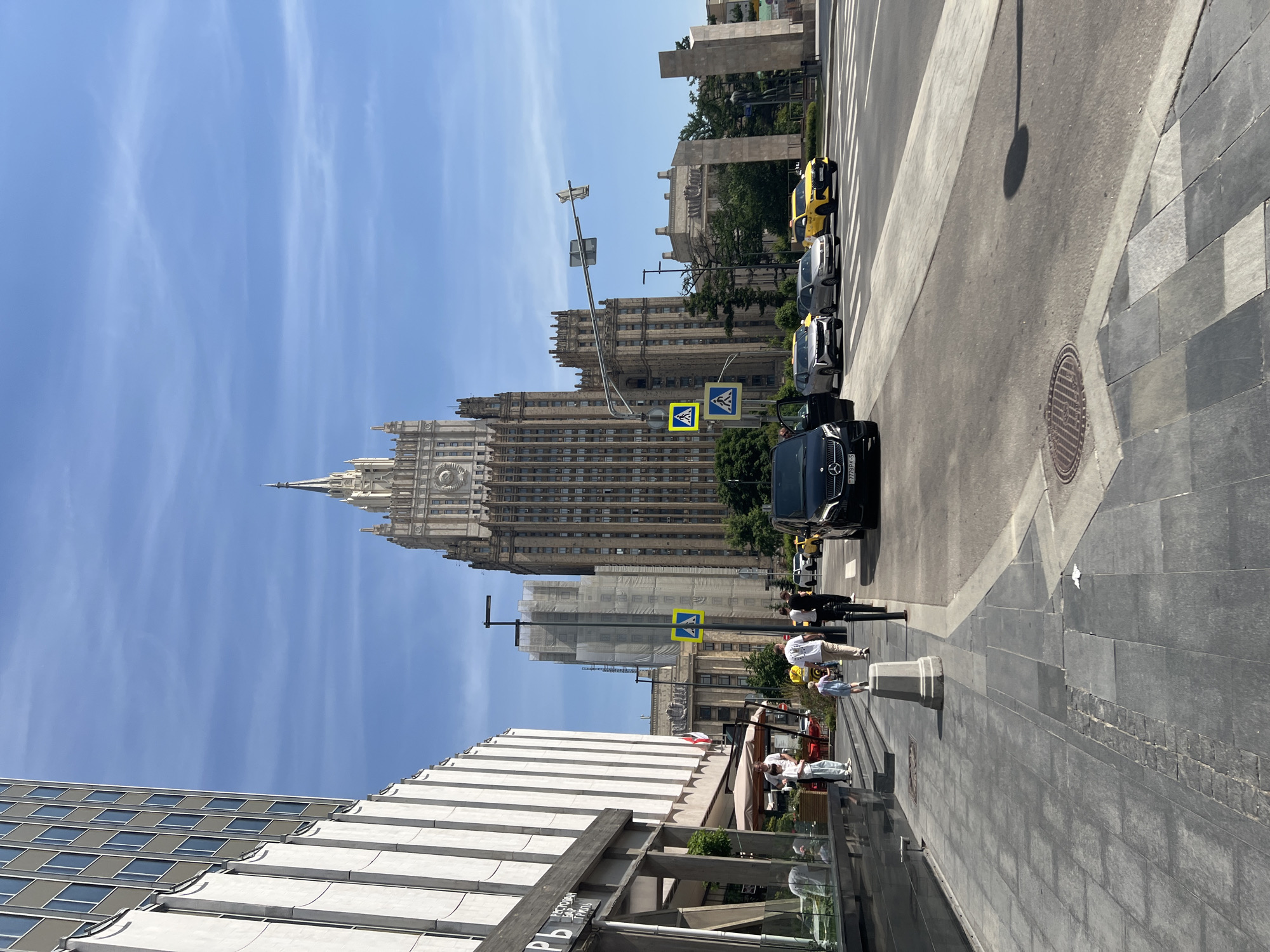 This image depicts a sunlit street scene in Moscow, Russia, prominently featuring the Main Building of the Ministry of Foreign Affairs on Smolenskaya-Sennaya Square in the background.
The Ministry of Foreign Affairs building is a large, multi-story Stalinist skyscraper with a central spire, capped by a star and a hammer and sickle emblem. Its facade is light-colored, with numerous rectangular windows and ornate architectural details.
In the foreground on the left, a modern building with a textured, light-colored vertical facade and rows of square windows rises. At its base, a commercial establishment, possibly a restaurant or cafe, features expansive glass windows and outdoor seating areas. Several individuals are visible on the adjacent sidewalk, some standing near the establishment, others walking. A child in a light blue outfit is walking away from the camera, followed by an adult in a white t-shirt and light trousers.
A multi-lane asphalt road occupies the middle ground. A clearly marked pedestrian crossing, indicated by white stripes on the road and two yellow square pedestrian crossing signs on a pole, spans the street. Multiple vehicles are present: a black Mercedes-Benz V-Class van (license plate 7779PK-5) is parked on the right side of the road with its front driver's side door open. Other vehicles on the road include a silver SUV, a light-colored sedan, and two yellow taxis (one SUV, one sedan), all traveling away from the viewer.
The sidewalk on the left is paved with grey rectangular tiles, with a concrete curb separating it from the road. A light-colored, bell-shaped bollard stands on the sidewalk. Further down the street, beyond the pedestrian crossing, a building undergoing construction or renovation is visible, covered in white scaffolding and netting. Trees and green foliage line parts of the street and background. On the far right, a sculptural or monumental structure composed of large, light-colored rectangular blocks is partially visible.
The sky is clear and blue with faint wispy clouds. The scene is brightly lit by sunlight, casting distinct shadows on the ground, notably a large oval shadow on the asphalt in the lower right.