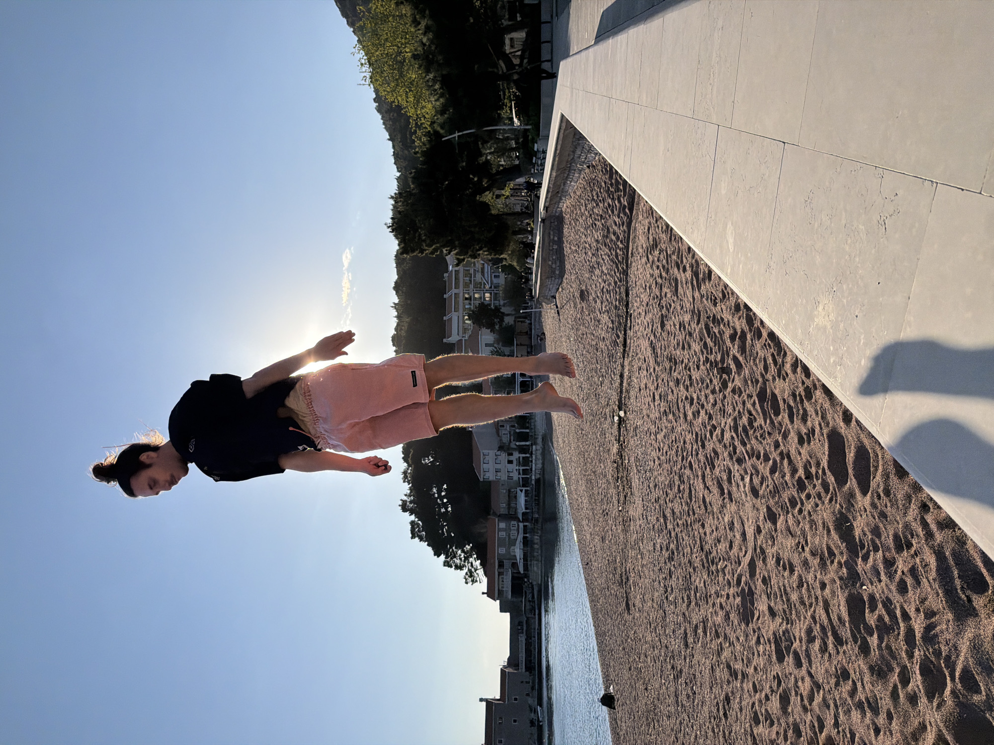 The image captures a person suspended in mid-air above a sandy beach, suggesting a jump or a deliberate floating pose for the camera. The individual, appearing male, is dressed in a dark short-sleeved top and light pink shorts, with their hair pulled back in a bun. They are barefoot, with arms slightly bent and hands open, and legs also bent, conveying a sense of weightlessness and movement.
The setting is a coastal area, specifically Petrovac centar, Montenegro. The scene is illuminated by the low-angled sun, positioned behind and to the left of the person, indicating either late afternoon or early evening. This strong backlighting creates a halo effect around the person's silhouette and casts long shadows across the beach and a bright, clear blue sky.
Below the person is a dark, coarse sandy beach featuring textured patterns and footprints. To the right, a clean, light-colored concrete wall or pathway extends diagonally, forming a clear boundary. Beyond the beach, calm water, likely the sea, stretches out, leading to a backdrop of white-colored buildings and houses typical of a coastal town. These structures are nestled at the base of lush green, tree-covered mountains or hills. The overall mood of the image is dynamic and serene. No discernible text is visible in the image.