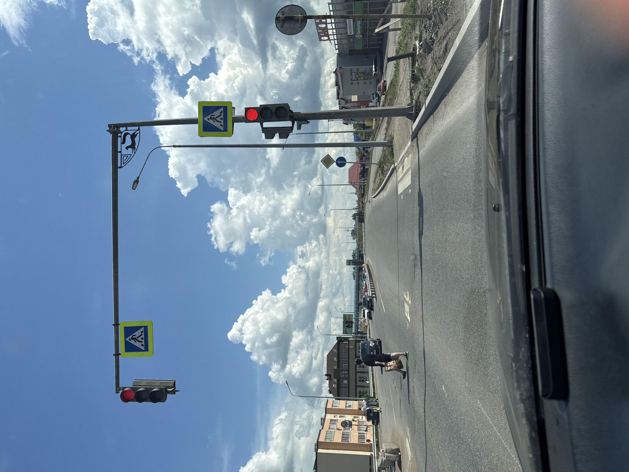 The image captures a street scene with a traffic light showing red, indicating a stop. A pedestrian crossing sign is visible above the traffic light. The road is paved, with a crosswalk marked on it, and several vehicles are present, some of which are moving. Buildings line the sides of the street, and trees and streetlights are also visible. The sky is blue with white clouds, suggesting fair weather. A person is walking across the crosswalk, carrying a bag. The scene appears to be in an urban or suburban area.
