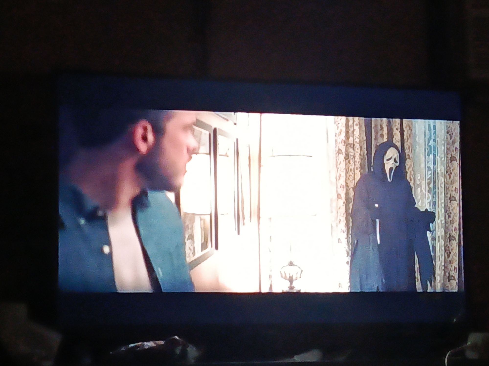 The image displays a scene from a screen, likely a TV. A man is visible on the left side of the screen, wearing a blue shirt that is open at the chest, and he is looking to the right. On the right side of the screen, there is a figure wearing the Ghostface mask and costume from the "Scream" movie franchise. Behind them are patterned curtains and a bright window. The background includes framed pictures on a wall and a small lamp. The image appears to be a photograph of a television screen displaying a scene from a movie or television show. There is no indication of any location, including Juan Griego, Venezuela.
