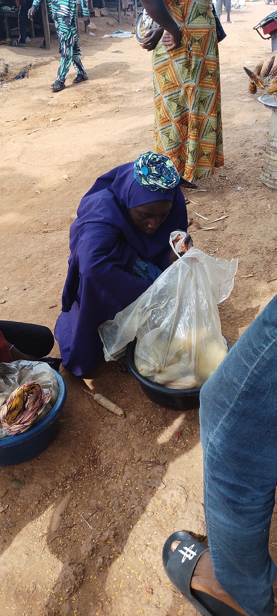 The image depicts a street market scene in Gumel, Nigeria. A woman wearing a dark blue hijab and a matching long dress is kneeling on the ground. She is interacting with a large plastic bag filled with light-colored items, possibly food. Her hands are inside the bag, and she appears to be handling its contents. Beside her, on the left, is a blue plastic bowl containing fabric items, possibly textiles or clothing. To the right of the woman, a black bowl filled with similar light-colored items is visible, also covered with a plastic bag. A single cob of corn lies on the dusty ground between the two bowls. In the background, other people are present, including one person wearing a vibrant green, black, and white patterned outfit, and another wearing a colorful, geometrically patterned skirt. A motorcycle is also visible in the background, suggesting a bustling market environment. The ground is made of dirt and scattered with small debris, consistent with an outdoor market setting. The overall impression is one of daily commerce and activity in the city.