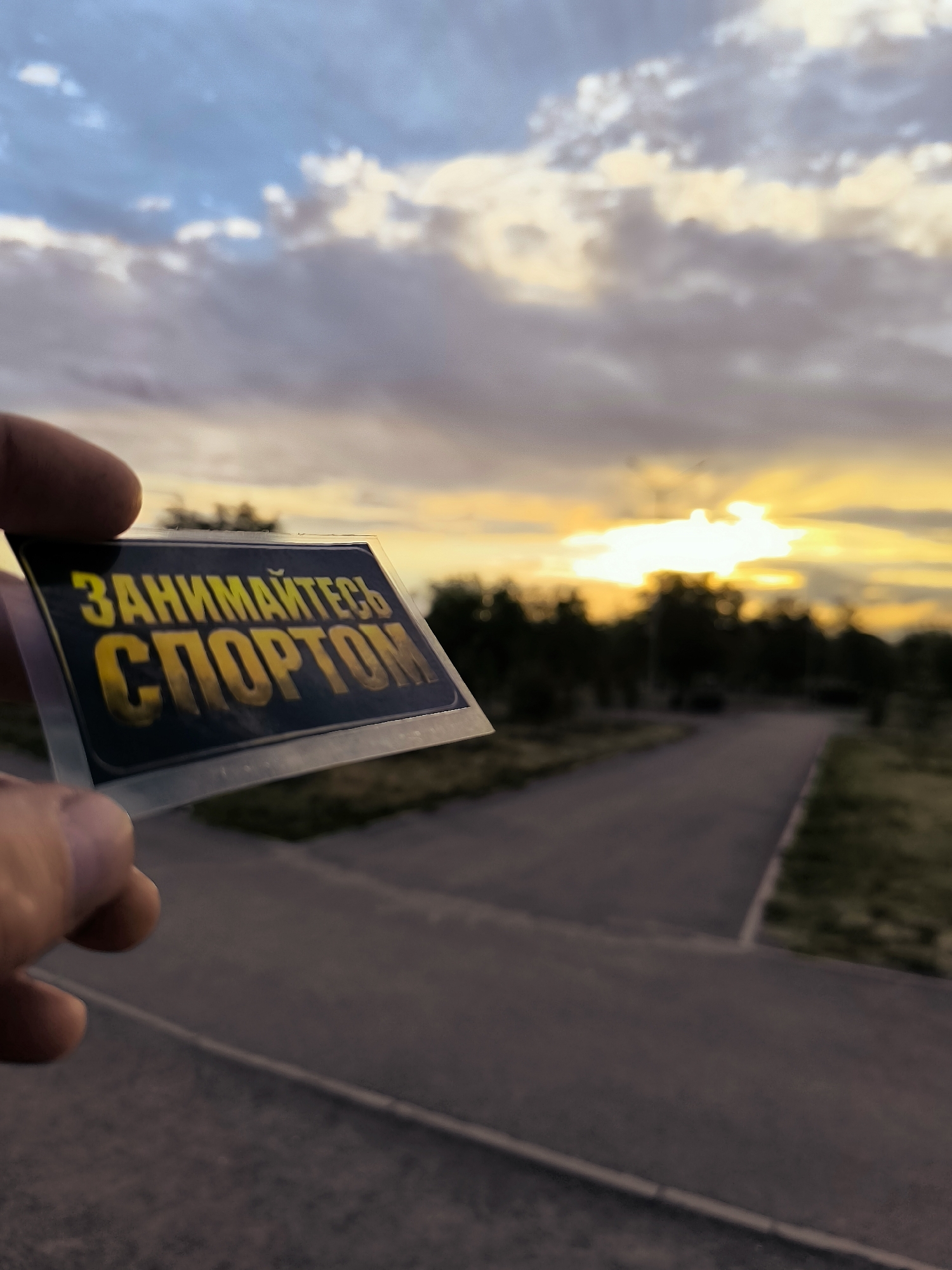 A close-up, eye-level shot features a sticker with yellow Russian text held by a person's thumb and forefinger against a blurred background of a sunset and a street. The sticker, placed at an angle to the camera, displays the text "ЗАНИМАЙТЕСЬ СПОРТОМ" in a bold, stylized font. The background shows a paved pathway or road curving to the right, bordered by grass and trees silhouetted against a bright, hazy sunset sky. Rays of sunlight pierce through dramatic clouds, casting a warm, orange and yellow glow over the scene. The lower portion of the image is dominated by the asphalt road, with some green patches of grass visible on the sides. The overall impression is of an outdoor setting during twilight.