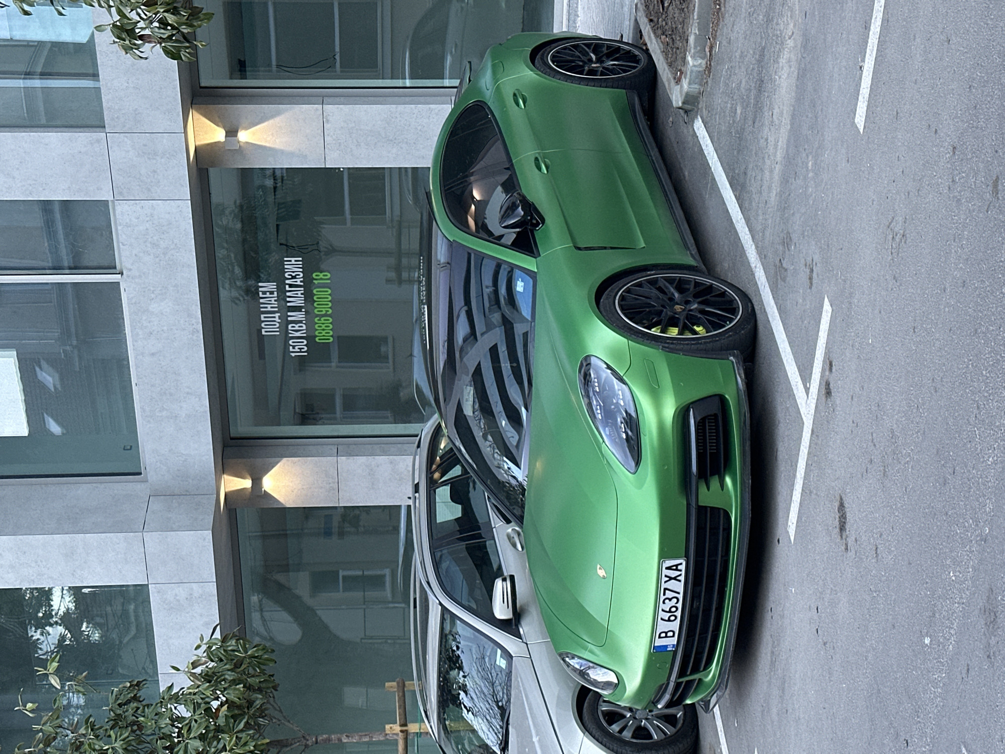 A green Porsche Panamera with license plate B 6637 XA is parked in front of a modern building. A white car is parked next to the Panamera. The building has glass windows. The words "ПОД НАЕМ 150 КВ.М. МАГАЗИН 0886 9000 18" are on one of the windows. The picture was taken on the street of Varna, Bulgaria.