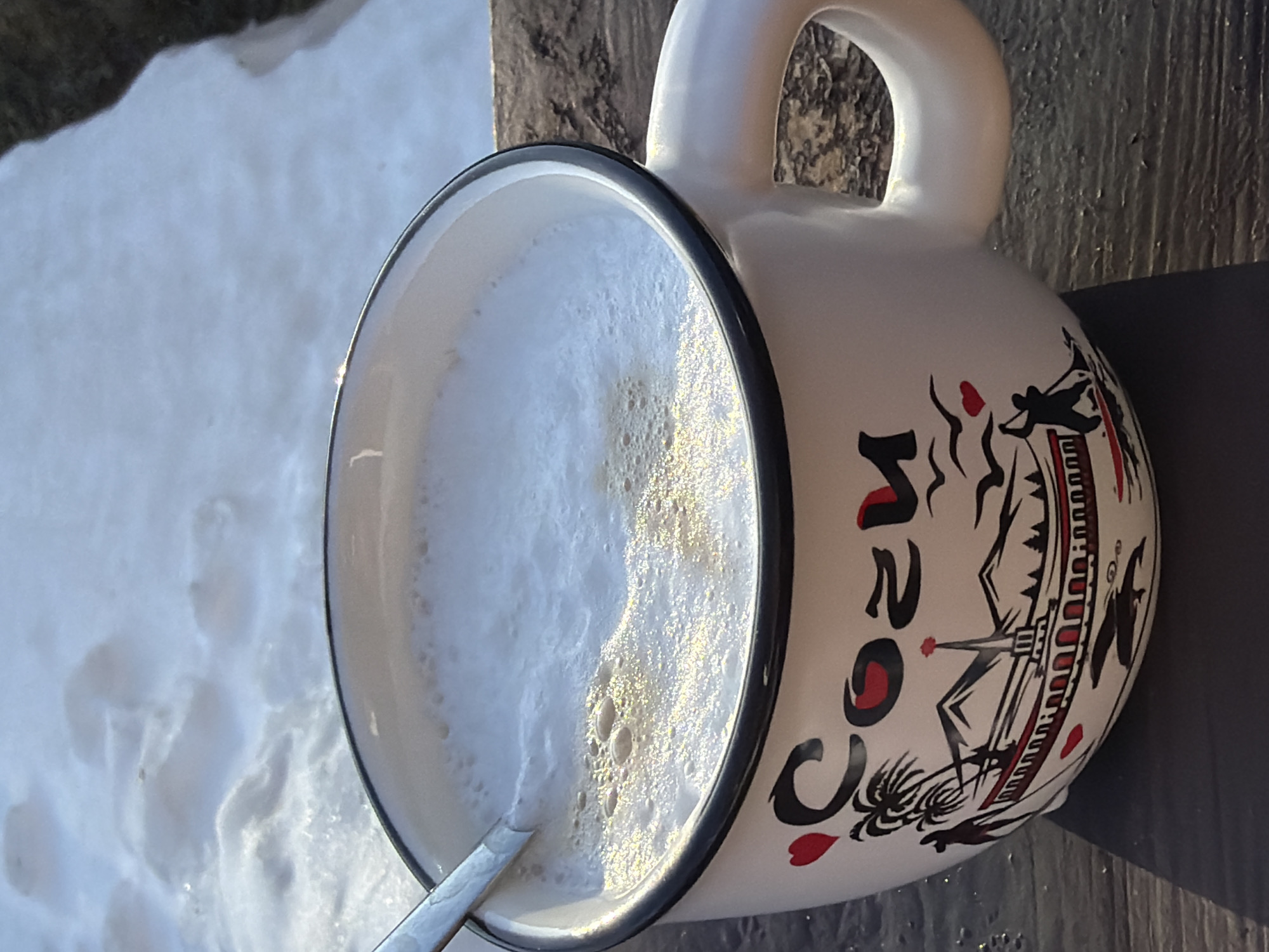 A high-angle, slightly tilted close-up shows a large mug filled with a frothy beverage, positioned on a weathered wooden surface outdoors. The mug is white with a black rim and handle, and decorated with a black and red illustration featuring palm trees, a stylized building, and the word "COKOJI". A metal spoon is partially submerged in the beverage. To the left, a patch of snow is visible, indicating a cold environment. The background suggests a natural setting with some vegetation partially visible. The location is Chaikovsky, Russia.