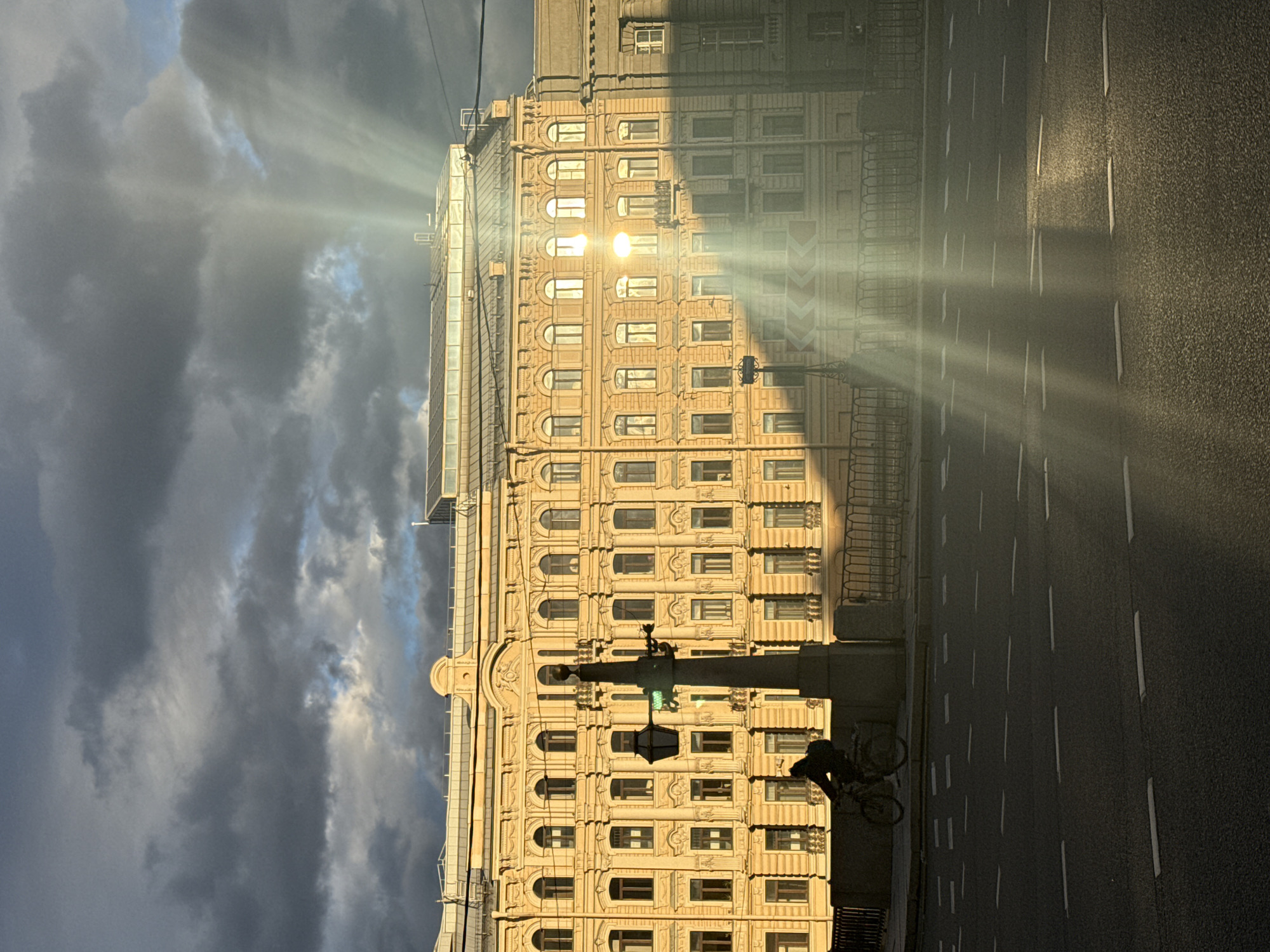 The image depicts a street scene in Saint Petersburg, Russia, captured during a late afternoon or early evening when the sun is low in the sky. The light casts long shadows and creates dramatic rays that pierce through some clouds and illuminate the facade of a grand, historic building. The building, likely an apartment block or office building, features ornate architectural details, including numerous arched windows and decorative stonework.
In the foreground, a street runs alongside the building, marked with white lane dividers. A lone cyclist is visible on the street, silhouetted against the light, suggesting they are either riding or have stopped. The sky is a mix of dark, dramatic storm clouds and patches of bright blue, hinting at changing weather conditions. The sunlight, breaking through the clouds, creates a luminous effect, bathing parts of the building and the street in a warm, golden glow, contrasting with the darker, shadowed areas.
The overall mood is one of dramatic beauty, with the interplay of light and shadow creating a sense of depth and atmosphere. The iconic architecture of Saint Petersburg is evident, with the building's style suggesting a historical period, possibly late 19th or early 20th century. There is no visible text in the image.