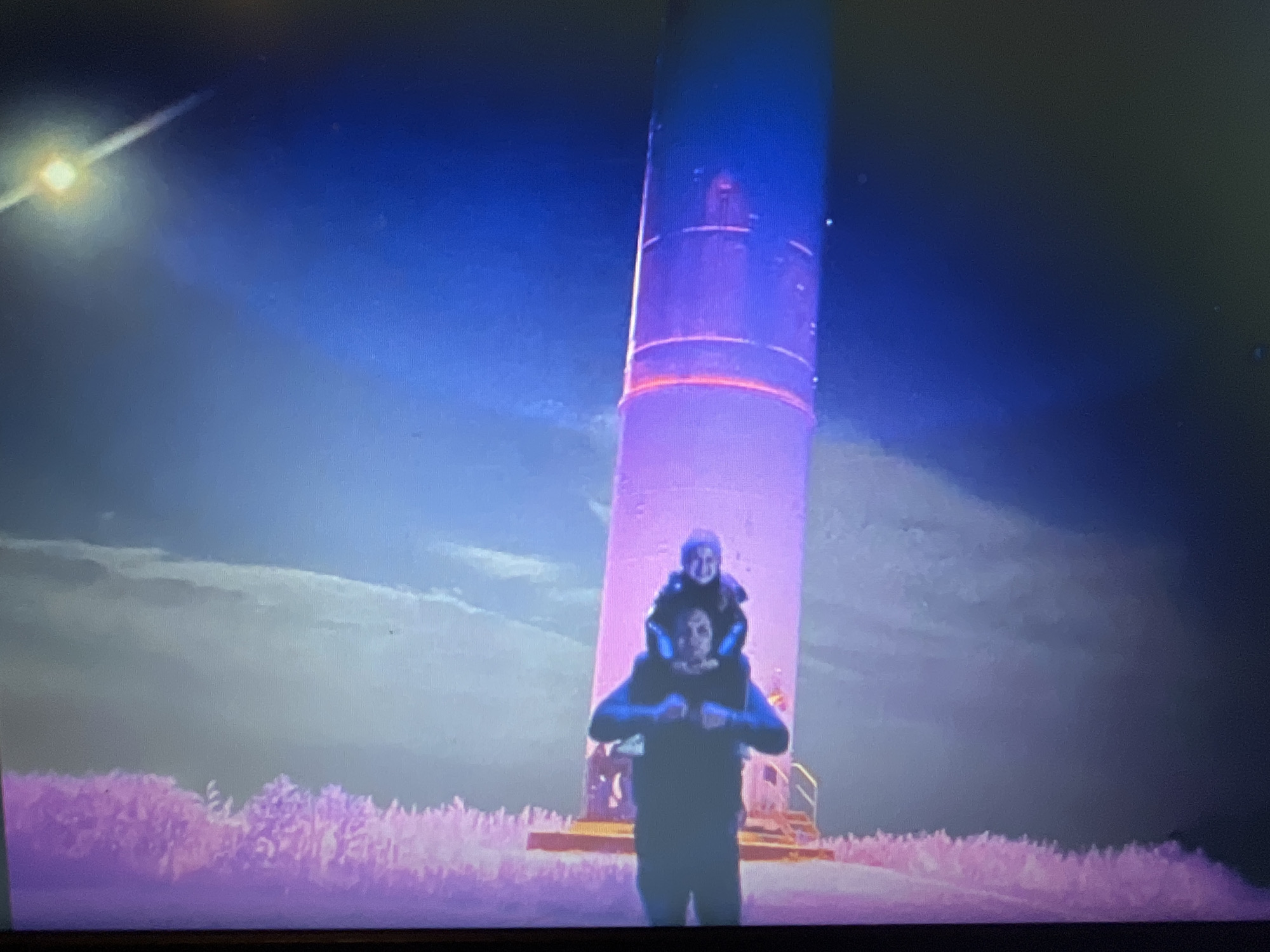 The image shows a man carrying a child on his shoulders in front of a large, illuminated tower. The man is wearing a dark jacket and the child is wearing a light-colored hooded garment. The tower is tall and cylindrical, with sections lit in purple and pink. There are clouds in the sky, and the landscape in the foreground appears to be a field of pinkish vegetation. The overall scene is dimly lit, with a bright light source visible in the upper left corner. The location is identified as Bilenke, Ukraine.