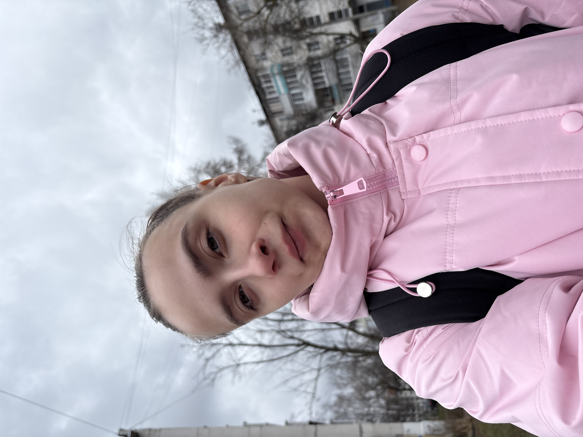 A low-angle, close-up portrait captures a young person, likely female, looking directly into the camera with a neutral to slightly contemplative expression. Her dark hair is pulled back, and she is wearing a vibrant light pink puffer jacket. The jacket features a prominent pink zipper, matching pink snap buttons on a placket, and a pink elastic drawstring with a silver stopper on what appears to be a sleeve cuff. A black inner layer or garment is visible around her neck and shoulders, along with black straps, possibly from a backpack.
The setting is outdoors under an overcast sky, suggesting cloudy daylight conditions. Bare tree branches are visible in the background, indicating a colder season such as late autumn, winter, or early spring. A multi-story apartment building with numerous windows is discernible in the blurred background, characteristic of a residential area. This scene is located in Chaikovsky, Russia. No visible text is present in the image.