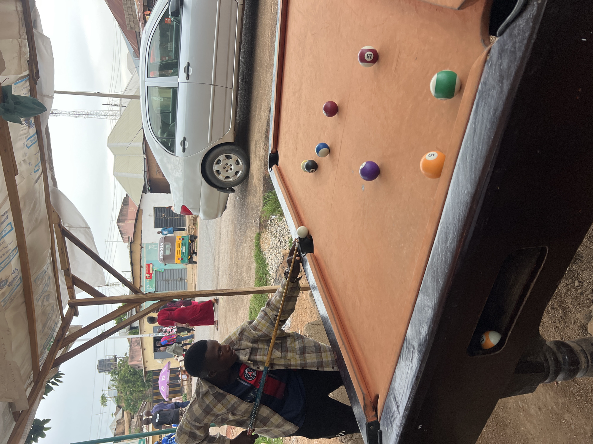 A man in a plaid shirt prepares to shoot a white cue ball on a pool table under a makeshift awning in Jos, Nigeria. The pool table is orange with several billiard balls scattered across its surface, including 5, 15, and a striped ball in the pocket. Behind the pool table is a silver car parked on the street. People and a vehicle marked "PTA-994" are visible in the background of the scene, indicating a public area within Jos. The environment is urban, with buildings and street activity, suggesting this is a local gathering spot.