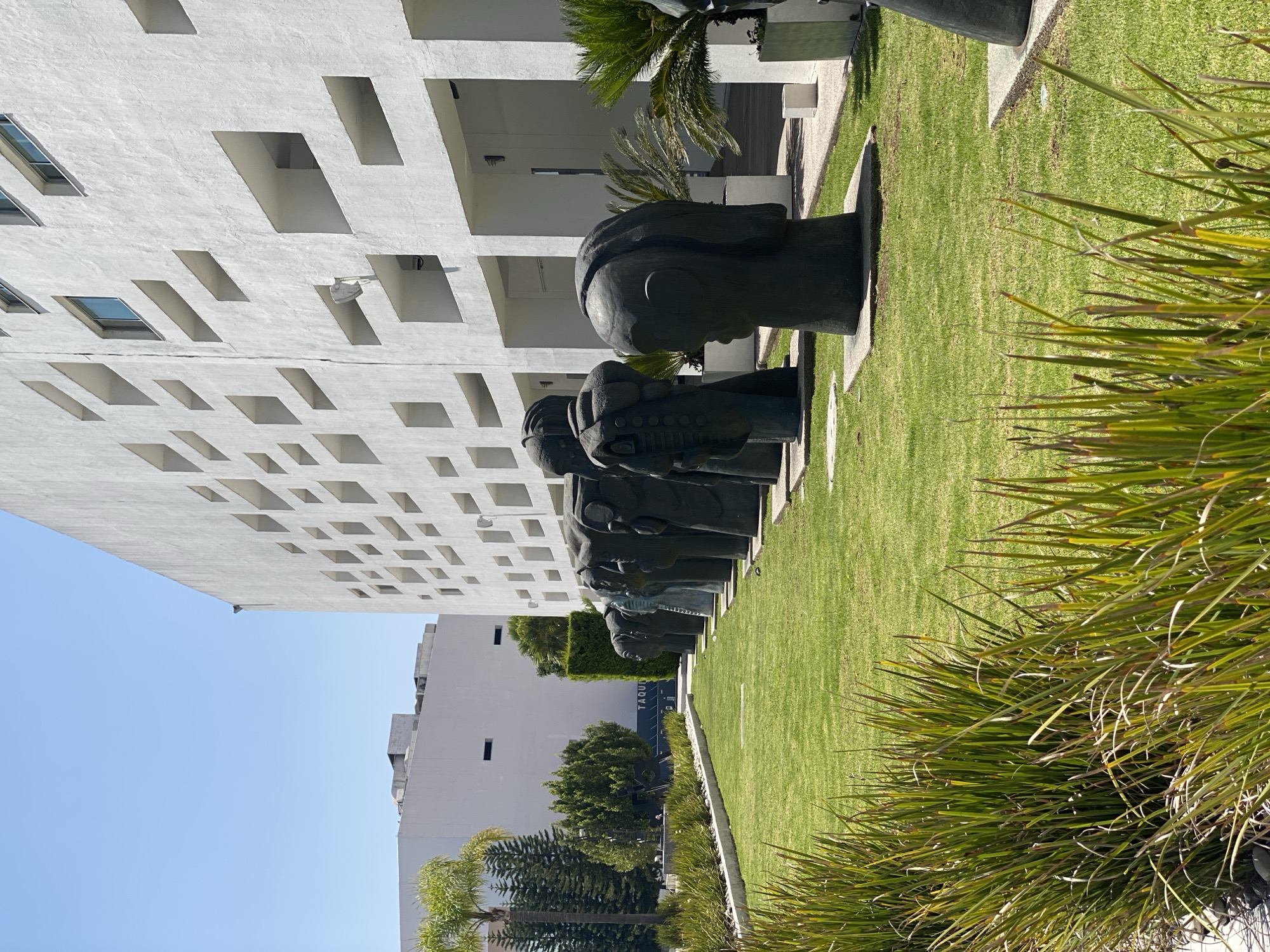 The image shows a row of head sculptures in a garden setting, with modern architecture in the background. The building is white with a patterned facade of square and rectangular cutouts. The sculptures, arranged on a grassy area, appear to be made of a dark metal or stone, each resting on a concrete base. Tall grasses and a palm tree frame the right side of the image, adding depth and texture to the scene. The clear blue sky suggests it is daytime. The location is Unknown, Unknown.
