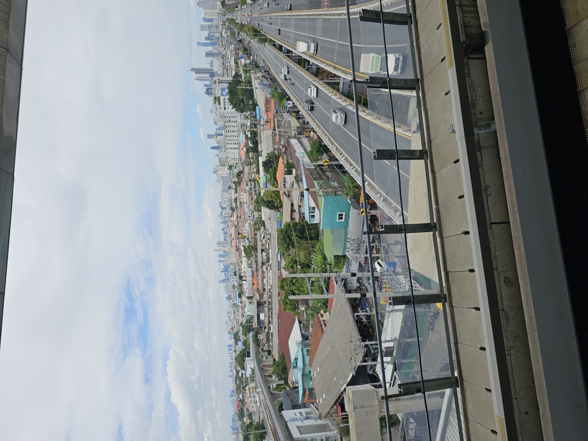 The image depicts an urban landscape in Bangkok, Thailand, characterized by a mix of residential buildings, commercial structures, and elevated infrastructure. In the foreground, a modern train track and platform are visible, suggesting a public transportation system. Beyond this, a sprawling cityscape unfolds, with numerous buildings of varying heights and architectural styles. A multi-lane highway runs through the scene, with vehicles including cars and trucks navigating its elevated sections. The sky is partially cloudy, indicating daytime. The overall impression is one of a densely populated and developed metropolitan area.