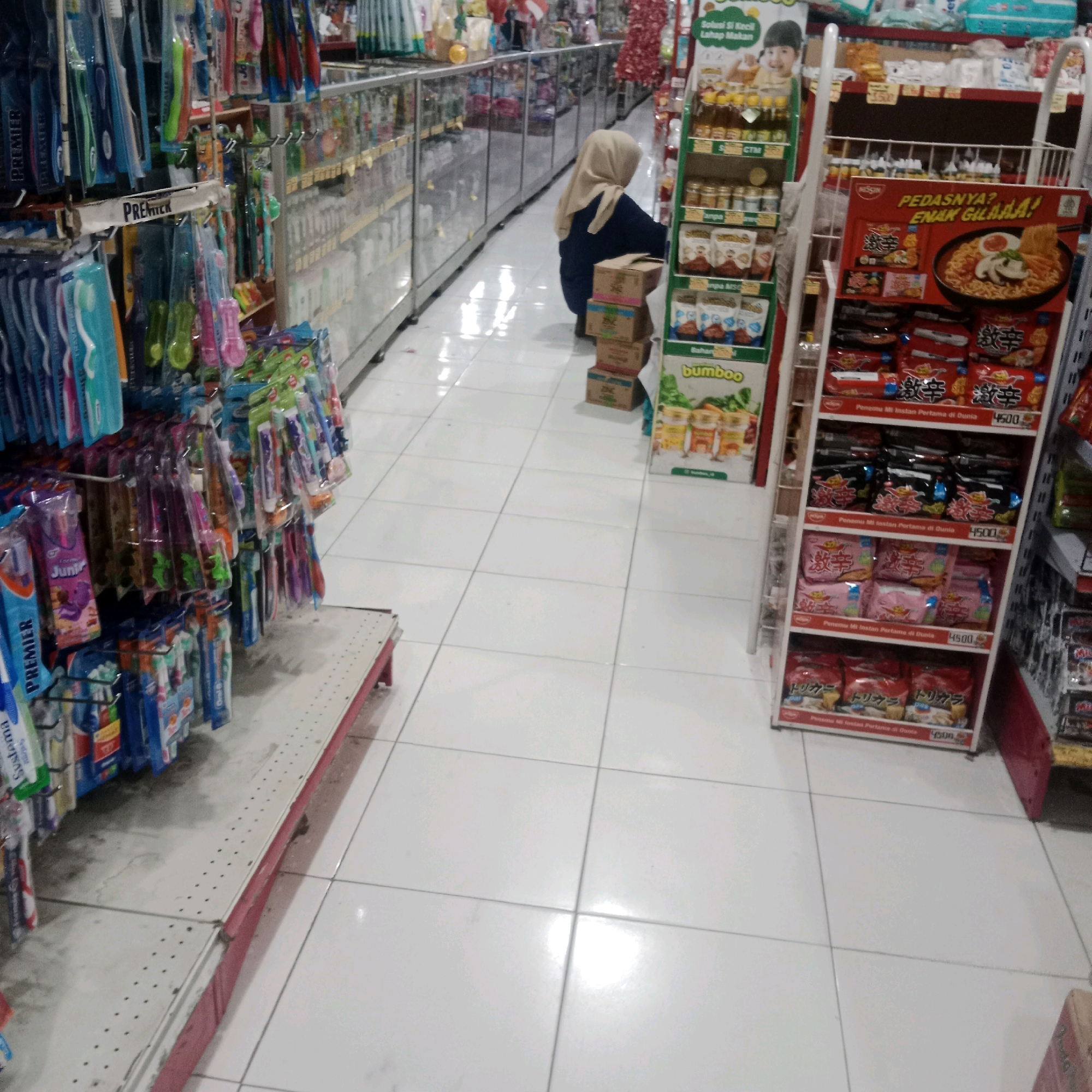 This image depicts an interior view of a retail store, possibly a minimarket or supermarket, in Tegowanu Wetan, Indonesia. The scene is brightly lit with white tiled flooring.
In the foreground to the left, a display shelf is laden with various toothbrushes, packaged in colorful plastic. These toothbrushes are hanging on hooks, suggesting they are organized for sale. Further to the right, a woman wearing a hijab and a dark top is crouched down, facing away from the camera. She appears to be interacting with merchandise on a lower shelf of a display unit. This display unit holds several stacked cardboard boxes.
The background features additional aisles of products, with shelves stocked with various consumer goods. On the right side of the image, a prominent display rack showcases packaged instant noodles, with a large promotional sign featuring a bowl of noodles. Other products are visible on this rack, including smaller packaged food items. The overall impression is that of a well-stocked retail environment.