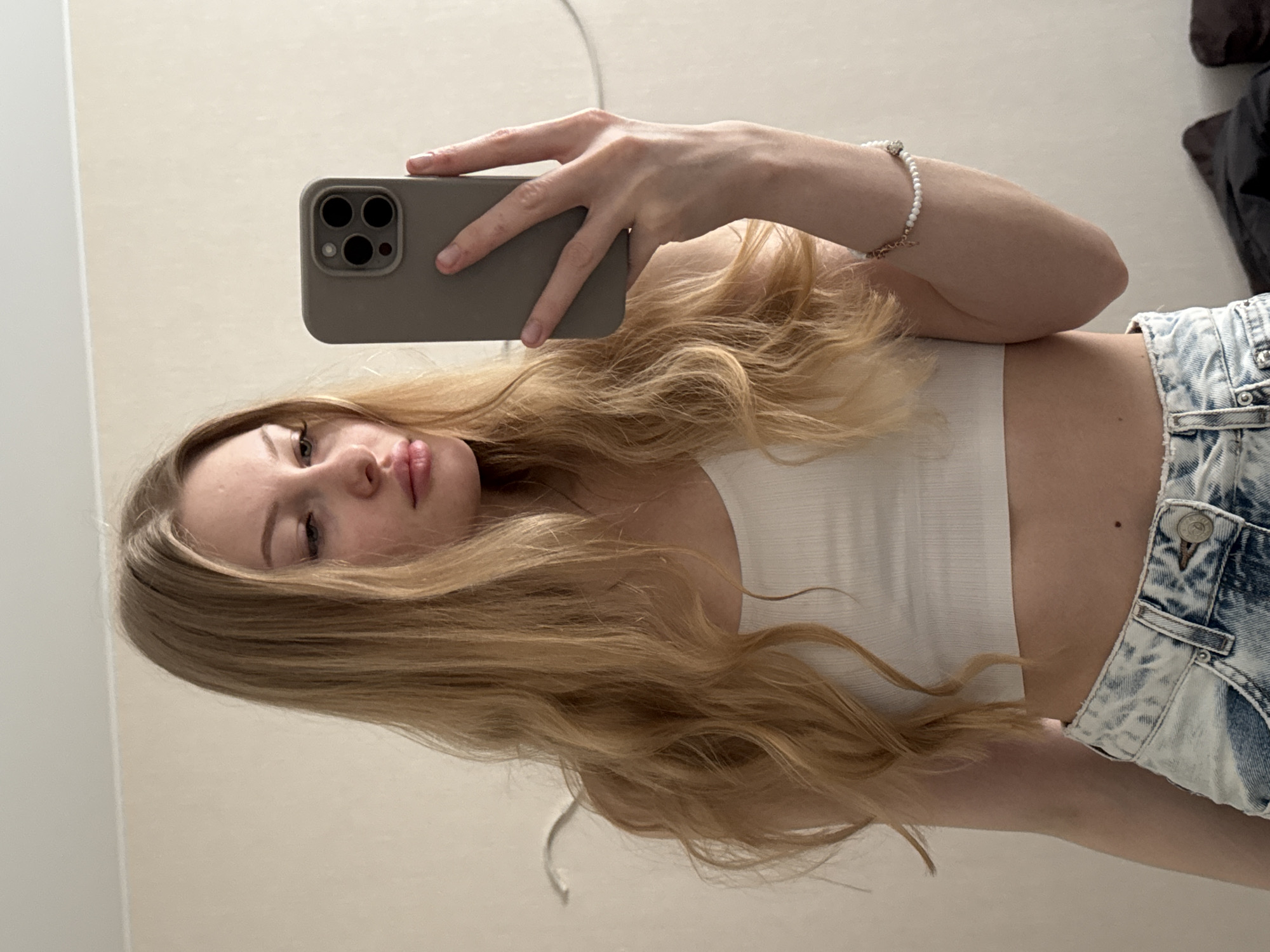 A young woman is taking a selfie in front of a pale wall. She is holding a smartphone with a triple-lens camera and a grey case in her right hand. She has long, wavy blonde hair. She is wearing a white tank top and light-wash denim shorts with visible wear patterns. A white beaded bracelet is on her wrist. Part of a black garment is visible in the upper right corner. There is no evidence suggesting this image was taken in Perm, Russia.