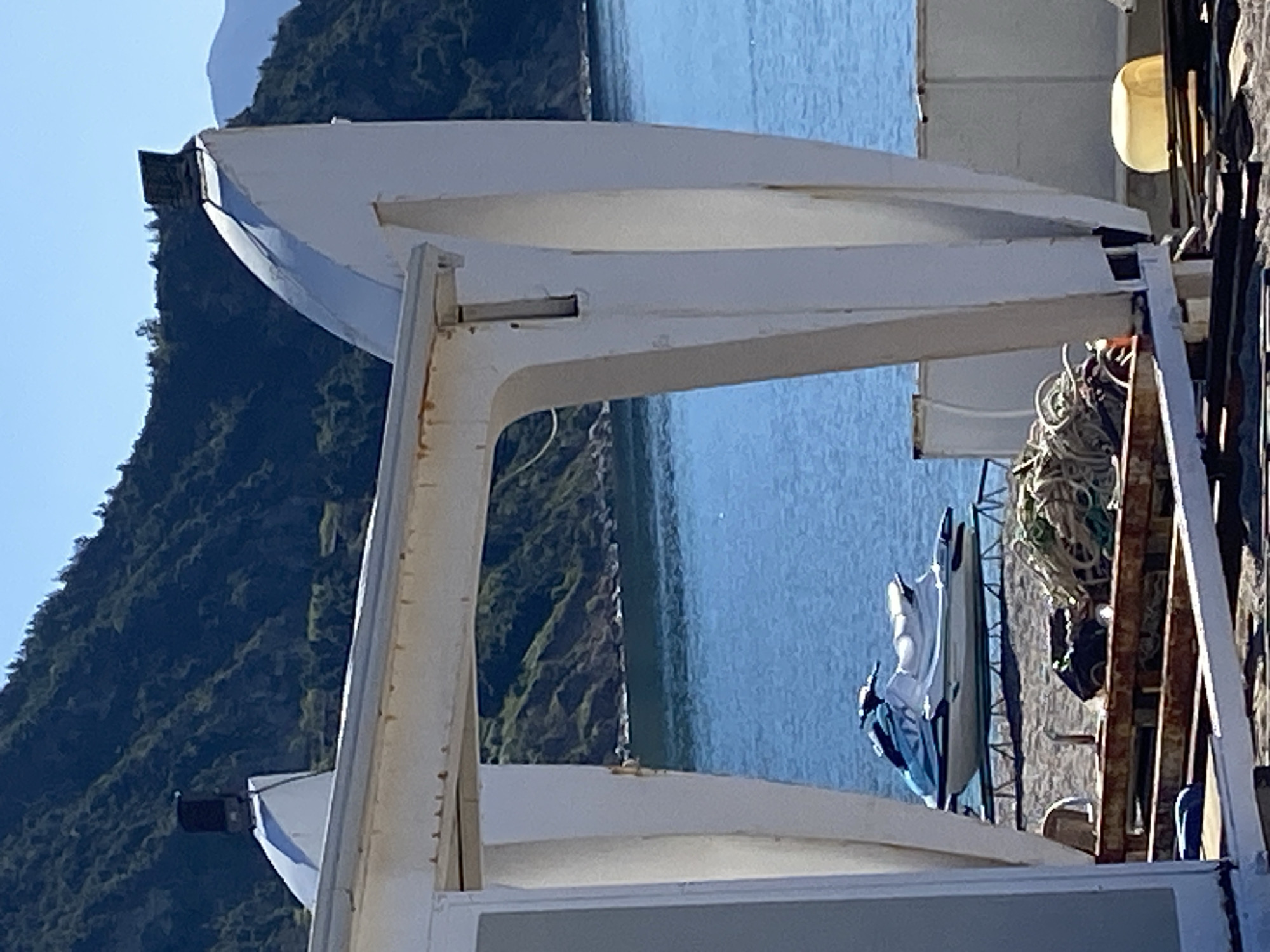 The image shows a boat deck in Prijevor, Montenegro. Two white lifeboats are stacked on a white metal frame on the side of the boat. Across from the boats on the dock, a jet ski, ropes, and rusted ladder can be seen. The background shows a body of water and green mountains.