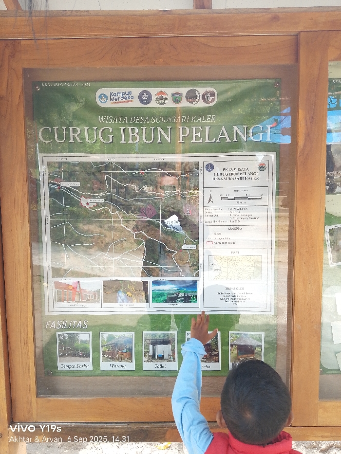 A rectangular, vertically oriented information board, enclosed in a light brown wooden frame and protected by a transparent sheet, is depicted at eye-level. The board details "WISATA DESA SUKASARI KALER CURUG IBUN PELANGI" (Tourism Sukasari Kaler Village Ibun Pelangi Waterfall). This location, Sukasari Kaler Village, is situated within the broader region of Sukadana, Indonesia.
The top section features a green banner with the "KAMPUS MERDEKA" logo and other institutional emblems, alongside the text "KNT-INIWA-S.173-2024". The main title, "CURUG IBUN PELANGI," is prominently displayed below. The central area of the board presents a topographic map of the waterfall area, complete with grid lines, labels, and an adjacent legend titled "PETA WISATA CURUG IBUN PELANGI DESA SUKASARI KALER." Below the map, three illustrative photographs show a building, a waterfall, and a panoramic landscape. The bottom section, labeled "FASILITAS" (Facilities), includes four smaller images with corresponding captions: "Tempat Parkir" (Parking Area), "Warung" (Stall), "Toilet," and "Gazebo." Additional text in the lower right attributes the creation of the map under "DIBUAT OLEH."
A young child, observed from behind, is positioned in the foreground. The child wears a red outer garment over a light blue long-sleeved shirt. Their right hand is raised, fingers splayed, and is in direct contact with the transparent protective sheet, specifically over the image labeled "Gazebo" in the "FASILITAS" section. Reflections from the outdoor environment are visible on the clear sheet. The ground beneath the child is light-colored and appears to be gravel or packed earth. A digital overlay in the bottom left corner reads "vivo Y19s Akhtar & Arvan 6 Sep 2025, 14.31".