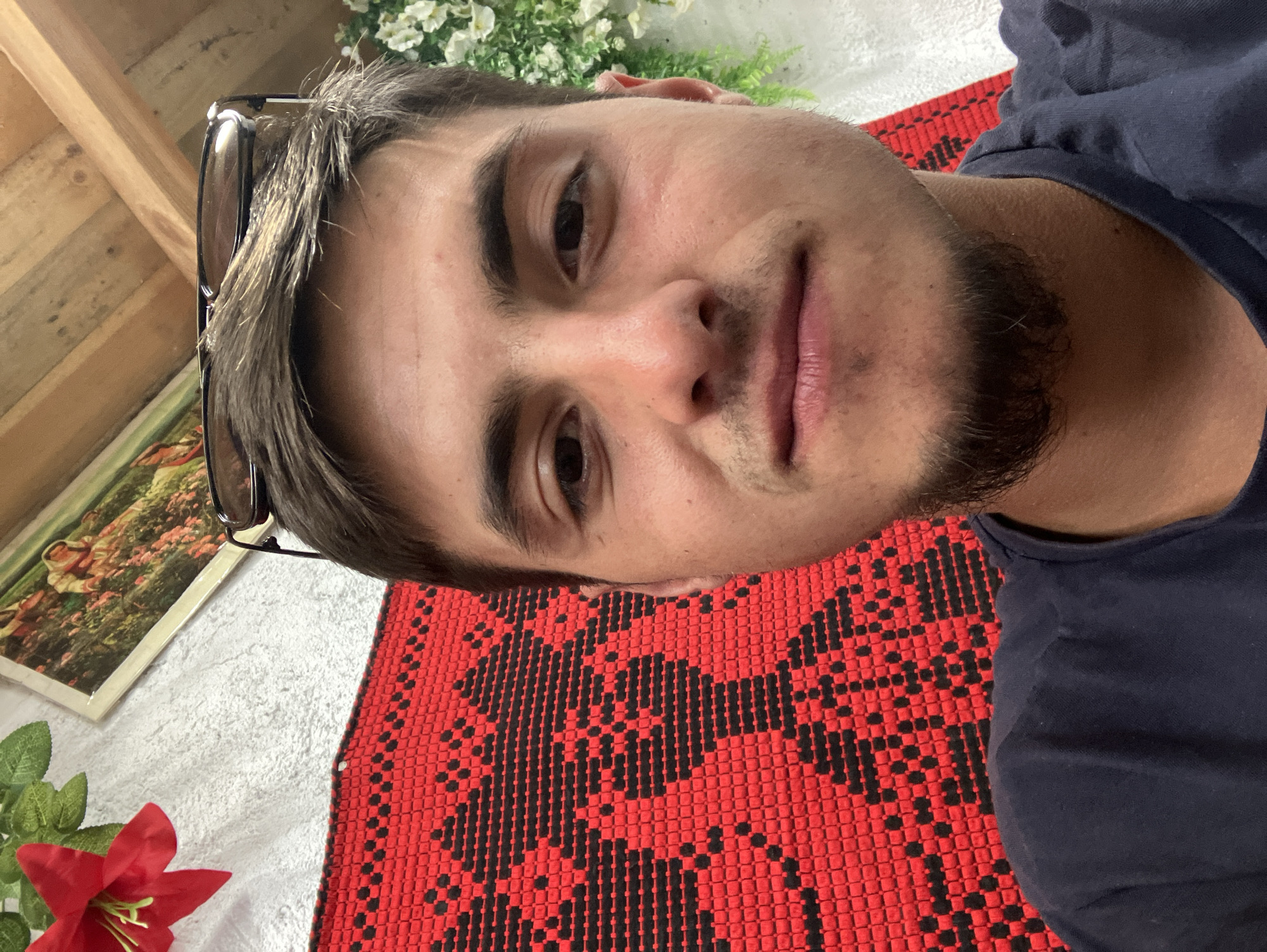 A medium close-up selfie of a Caucasian male with dark hair and brown eyes. He has a light beard. Sunglasses are perched on his head. He is wearing a dark blue t-shirt. The background includes a red and black patterned textile, possibly a rug or tablecloth, and white flowers are visible. There is also a framed picture behind him. I can not identify the location as the city of Komuniga, Bulgaria.
