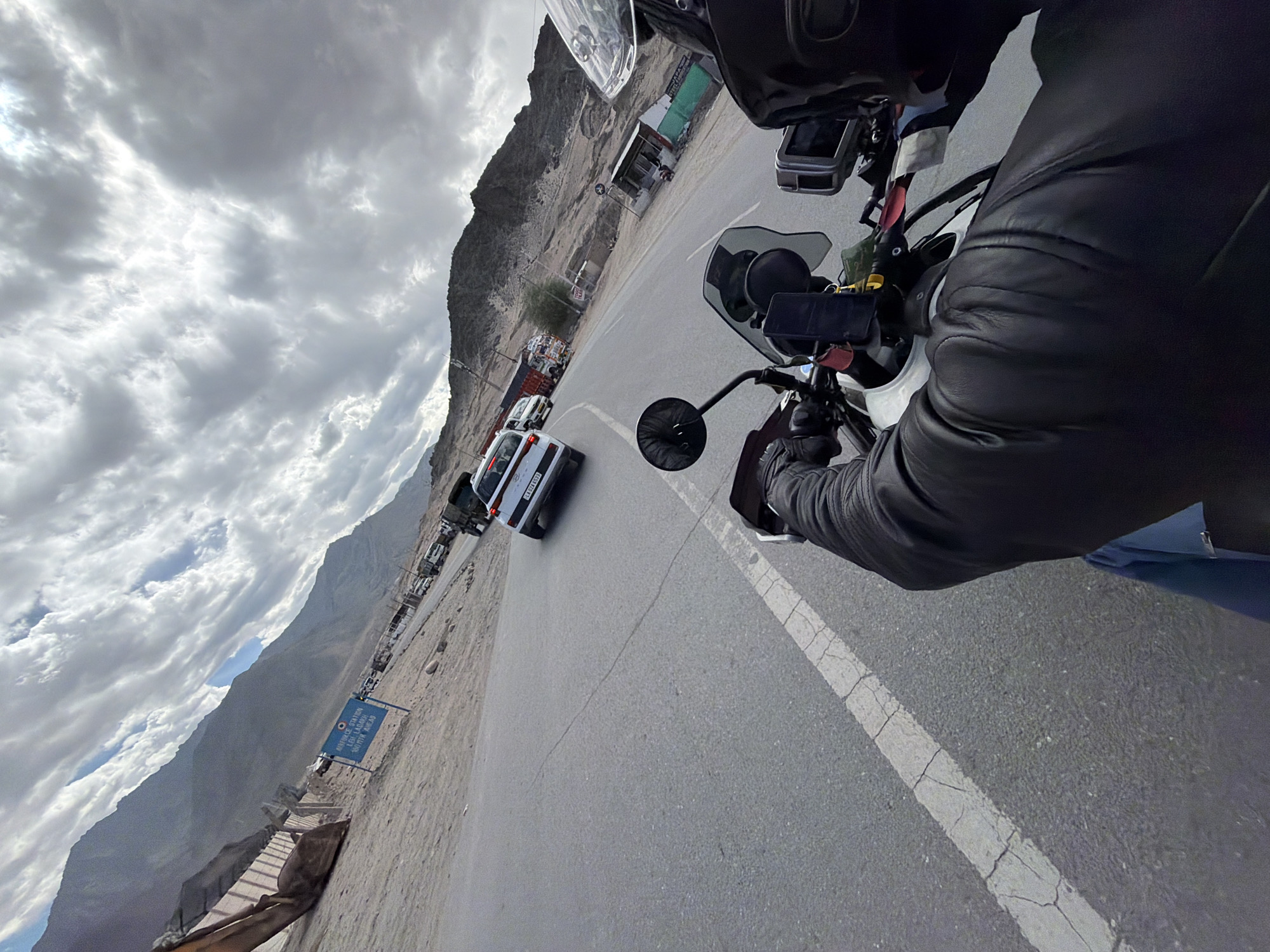 The image, captured from the perspective of a person riding a motorcycle, shows a road in Spituk, India. The road is paved and marked with a white line. In the distance, a white car is seen driving on the road, followed by several trucks and other vehicles. Mountains rise in the background under a cloudy sky. On the left side of the road, there is a sign that reads "AIRFORCE STATION LEH LADAKH 5 KM AHEAD." A construction vehicle is parked on the sandy terrain to the left of the road. The motorcycle rider is wearing a black leather jacket and a helmet. The overall scene depicts a journey on a road leading to an air force station.