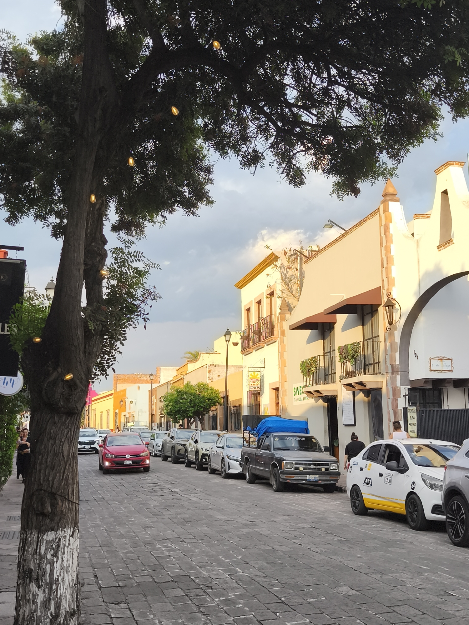 The image shows a street scene in Querétaro, Mexico. The cobblestone street is lined with parked cars, including a red Volkswagen Polo, a grey pickup truck with a blue tarp in the bed, and a white taxi with yellow stripes. A large tree stands on the left side of the frame, adorned with decorative string lights. On the right side of the street, buildings with architectural details are visible, including arches, balconies, and awnings. There are some people near the parked cars and a restaurant called SWEX. Signage is visible on some of the buildings, and there is a streetlamp further down the street. The sky is overcast with clouds.