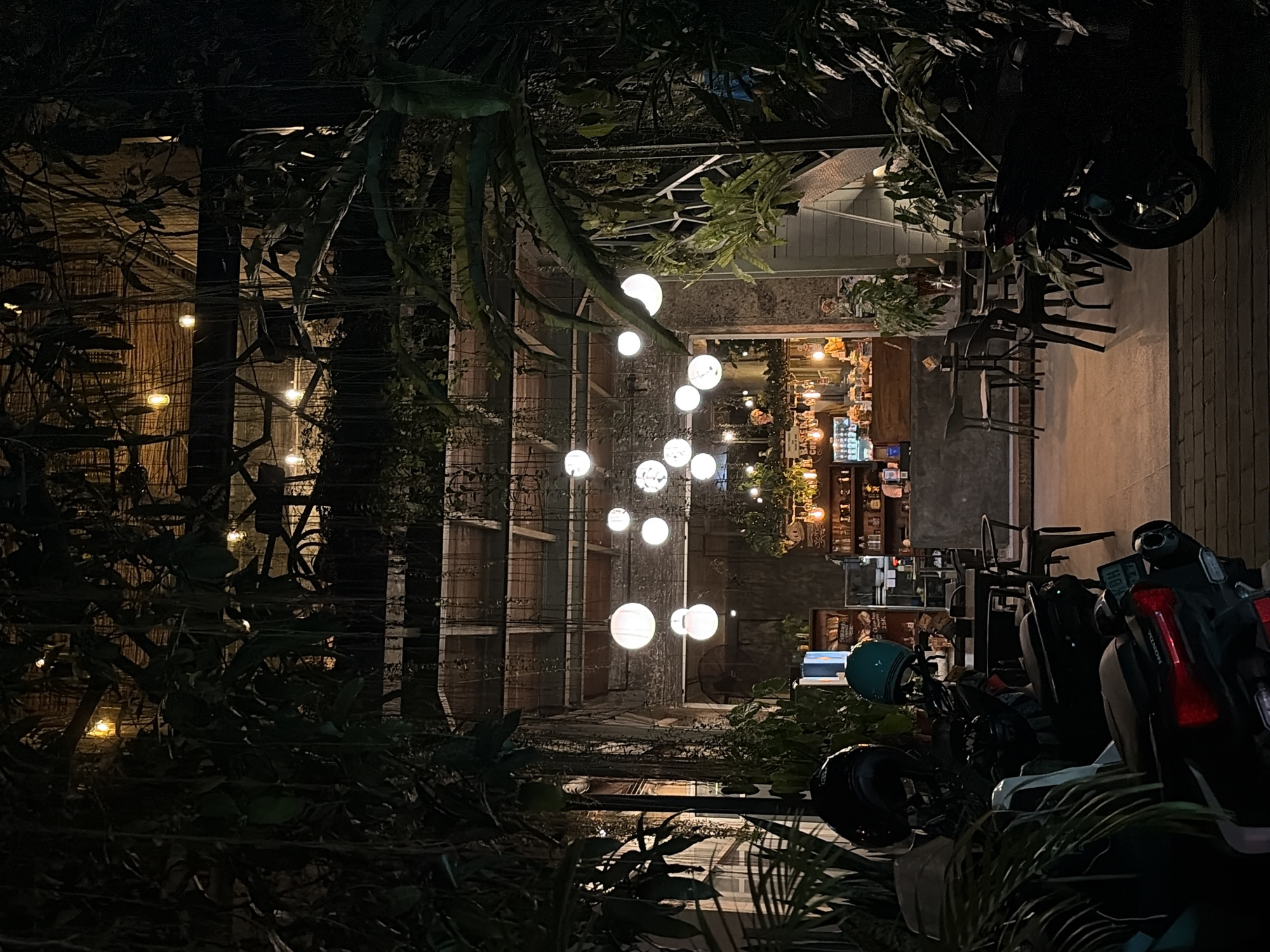 The image depicts an outdoor seating area of a cafe at night in Ubud District, Indonesia. Hanging plants and vines create a canopy over the scene, with string lights and spherical lanterns illuminating the space. In the foreground, several parked motorcycles are visible, some with helmets on the handlebars. Beyond the seating area, the interior of the cafe is visible, showing a counter with shelves stocked with bottles and drinks. The overall ambiance is dimly lit and tropical.