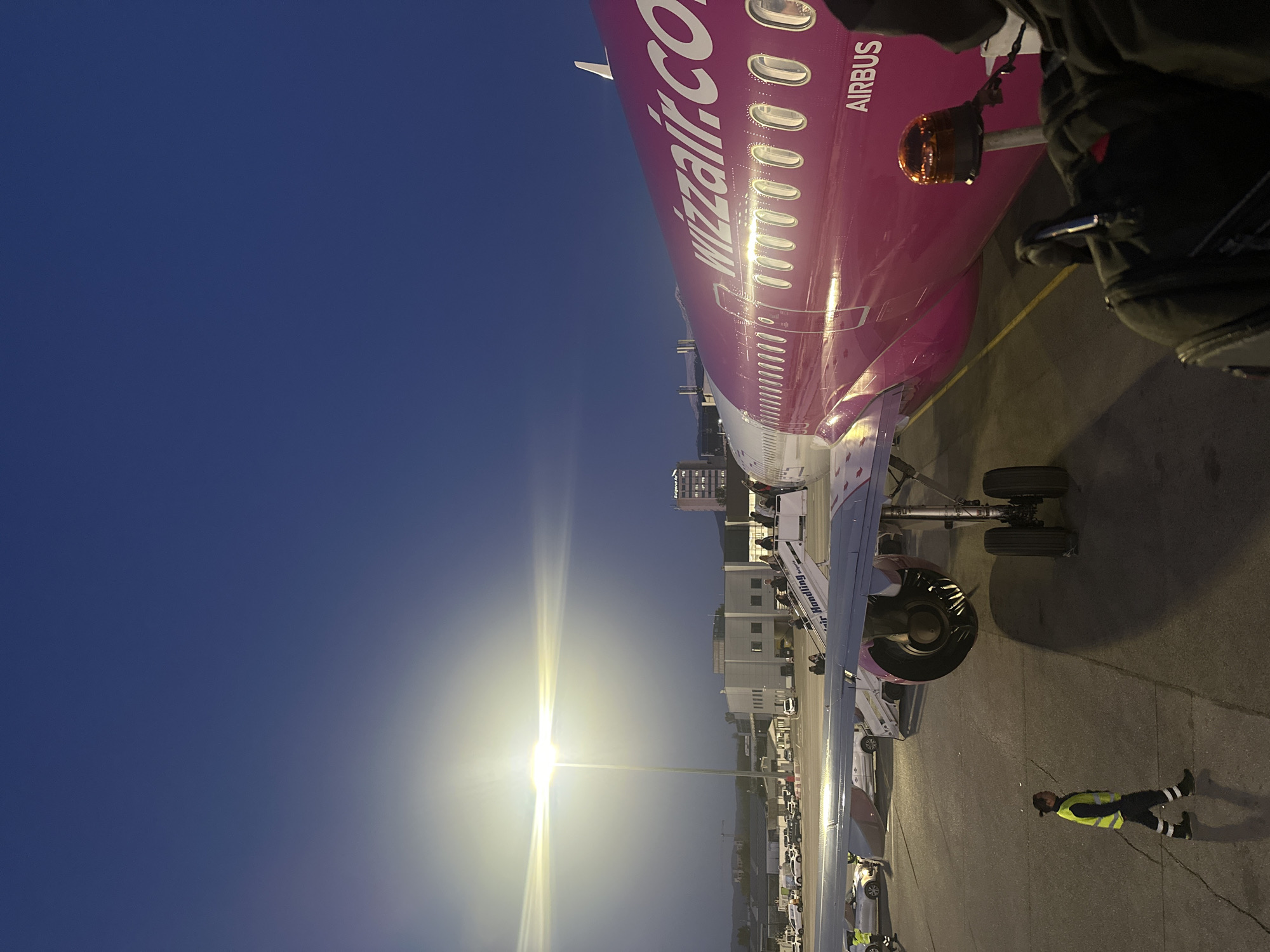 The image shows an Airbus aircraft with the Wizzair.com logo on its fuselage at Sofia Airport (SOF) in Sofia, Bulgaria. It is parked on the tarmac at night, as evidenced by the dark blue sky and artificial lighting. The aircraft's landing gear is visible. A ground crew member wearing a high-visibility vest is standing near the wingtip. The perspective is taken from inside the plane, possibly as passengers disembark. A bright light pole illuminates the scene. Buildings are visible in the background.
