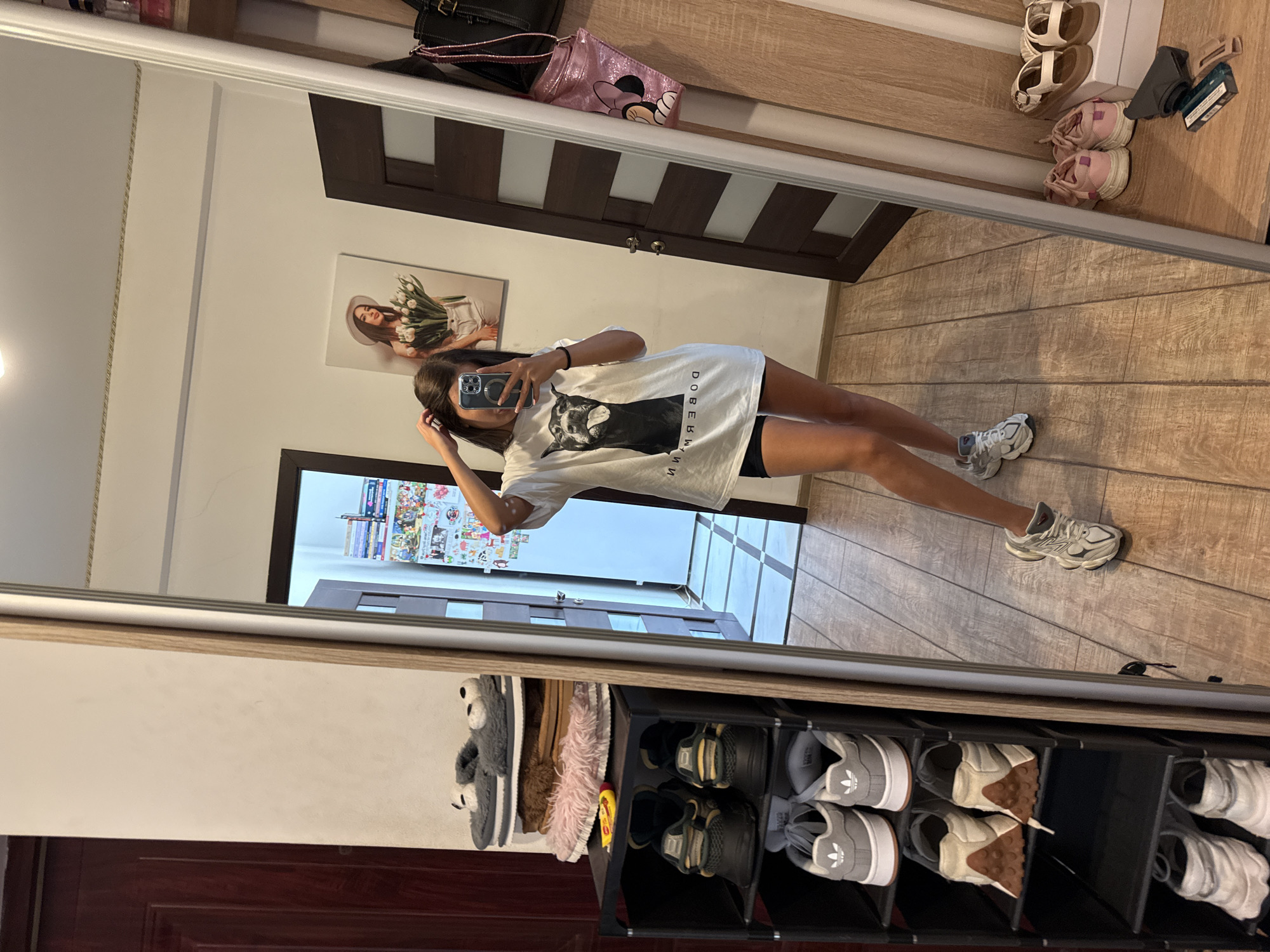 The image depicts a young woman taking a mirror selfie in what appears to be a bedroom or dressing area. She is wearing a white oversized t-shirt with a graphic of a Doberman on it and black shorts. In the background, a shoe rack is visible, filled with various pairs of sneakers and slippers. A large mirror reflects the woman and the room, including a door, a piece of art on the wall, and a glimpse of what might be a hallway or another room. The floor is made of wood-look tiles. There is no indication of the specific location in Chișinău, Moldova, beyond the presence of common household items.