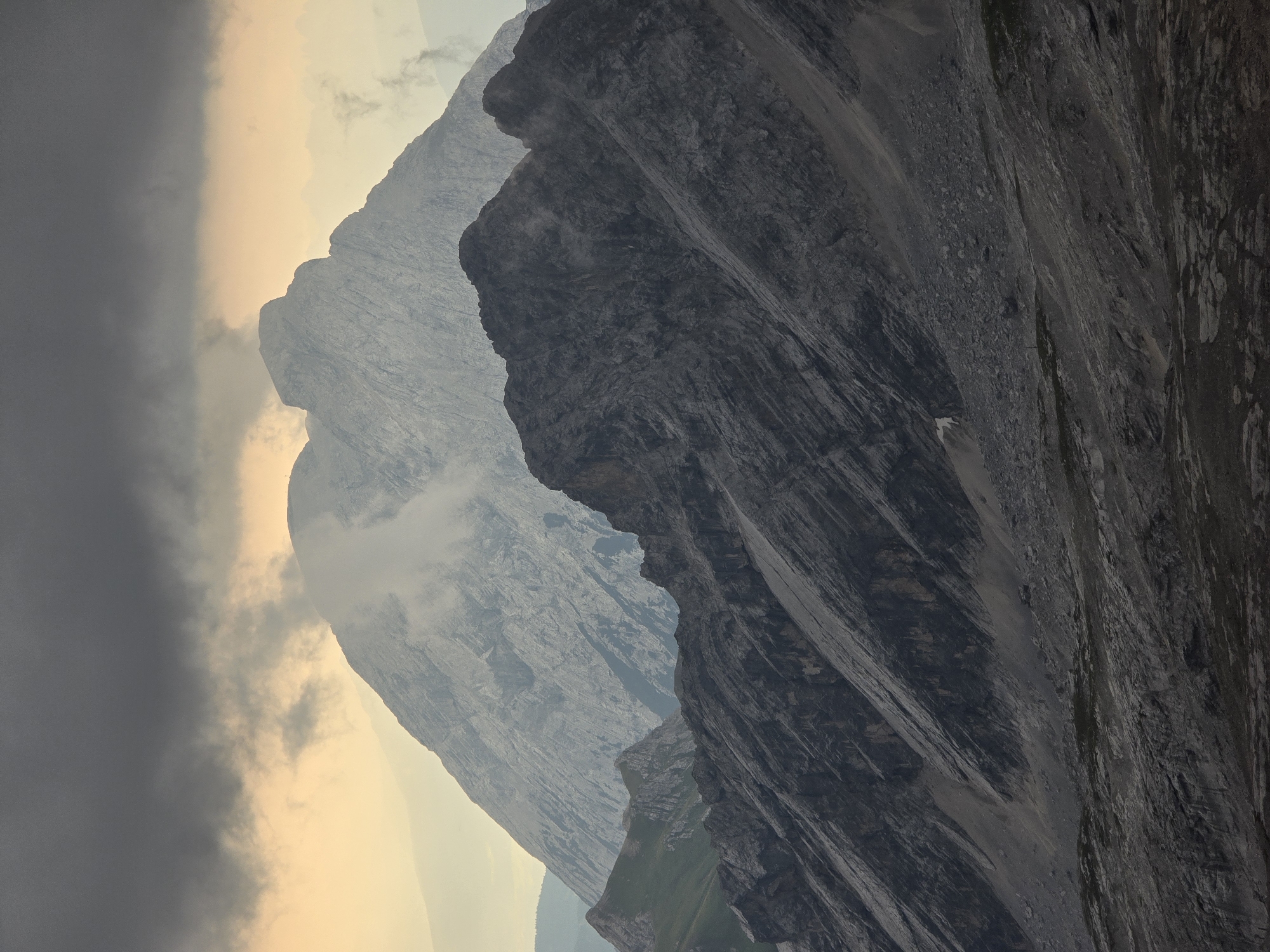 This image depicts a mountainous landscape, possibly near Garmisch-Partenkirchen, Germany, characterized by layered peaks under a dramatic sky. In the foreground, a dark, rugged mountain range with visible striations dominates the lower portion of the frame. Beyond this, a lighter, more expansive mountain peak emerges, partially obscured by clouds and mist, its slopes displaying intricate geological patterns. In the distance, a very faint, hazy mountain range is visible. The sky is a mix of dark, heavy clouds in the upper portion and a warmer, lighter hue near the horizon, suggesting either dawn or dusk. The overall scene conveys a sense of natural grandeur and a potentially overcast or changing weather condition in a high-altitude environment. There are no visible people or specific man-made objects.