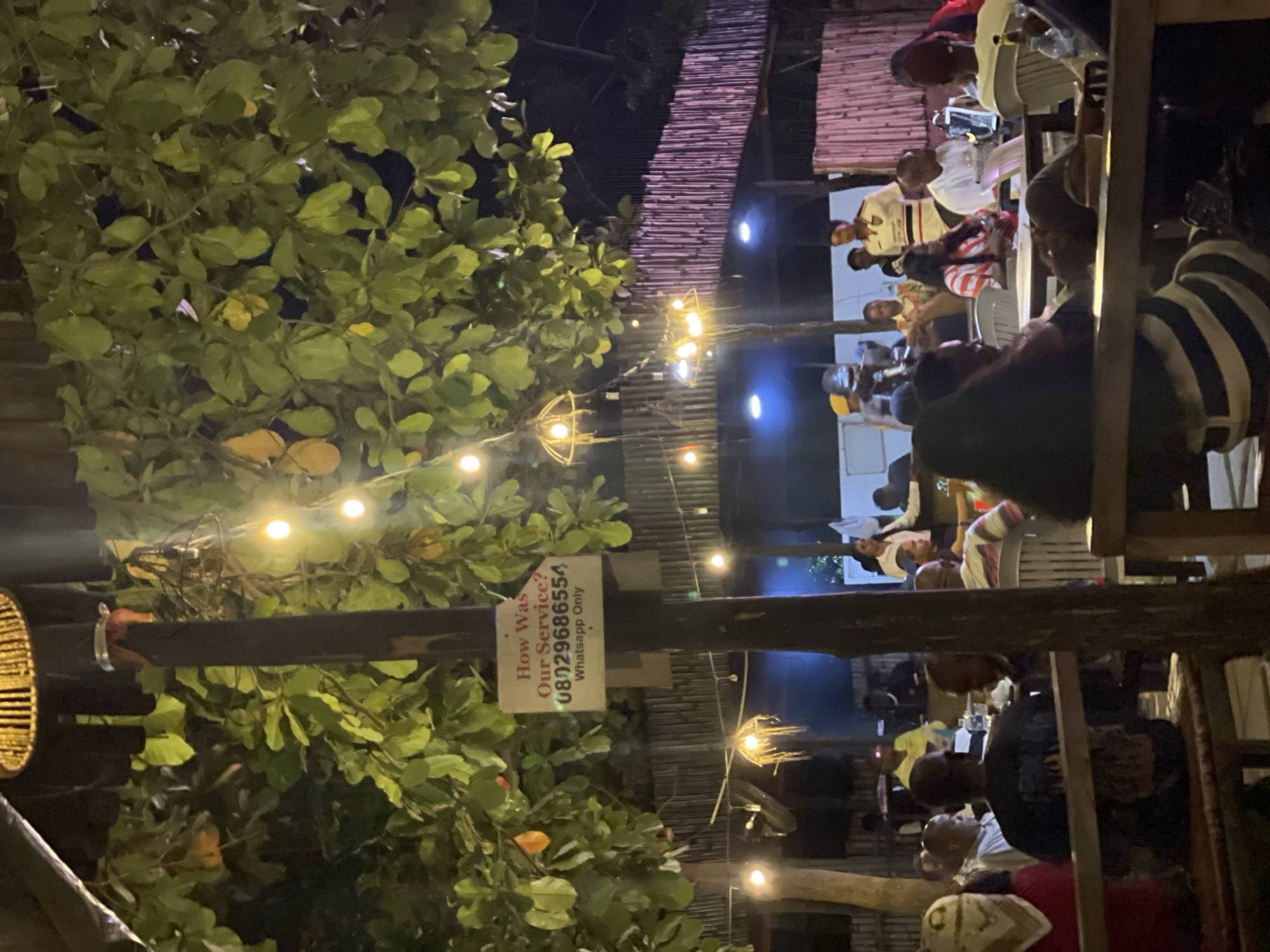 The image depicts an outdoor dining area, likely in Abuja, Nigeria. Patrons are seated at tables, illuminated by string lights. Lush foliage surrounds the area, and a sign asking about customer service is visible. People are seated and appear to be socializing.