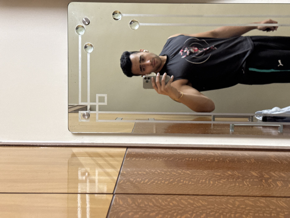 A man takes a mirror selfie. He wears a black t-shirt and dark pants with a green stripe. The mirror is rectangular with a decorative border featuring geometric designs and clear glass circles. It is mounted on a wall above a wooden dresser with varying shades and patterns. The man holds a smartphone, capturing his reflection with a focused gaze.