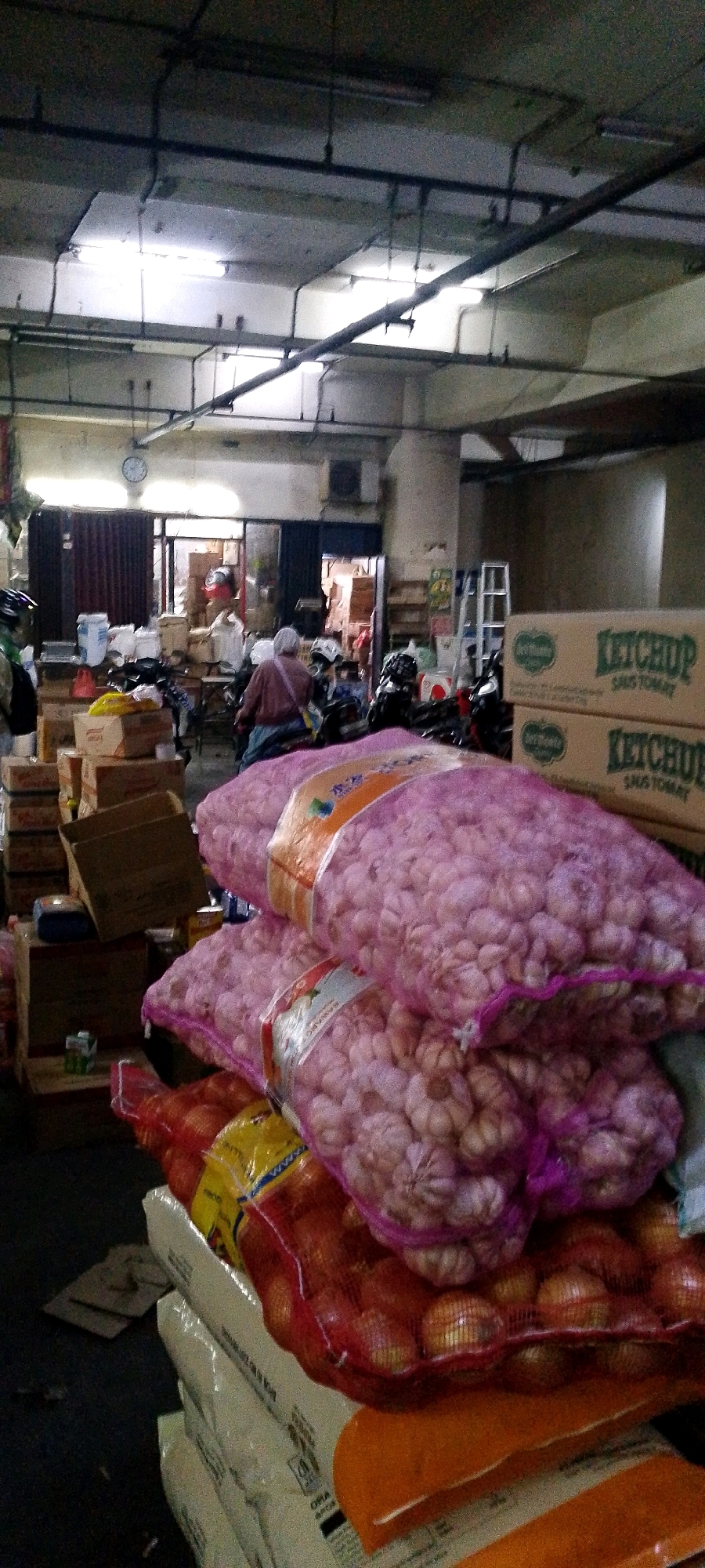 The image depicts an indoor market scene in Bandung City, Indonesia. In the foreground, large netted bags filled with what appear to be onions and garlic are stacked on top of each other. The bags are transparent, showing the produce within, and have labels attached. Behind the produce, there are several cardboard boxes, some stacked neatly and others piled haphazardly.
In the mid-ground, a person wearing a maroon top and a headscarf is seated on a scooter, facing away from the camera. Several other scooters are also visible, suggesting this is a place where motorized transport is common. More boxes and bags are scattered around this area. In the background, there are doorways leading to what appear to be shops or storage areas, with more boxes and shelves visible.
The environment suggests a commercial setting, possibly a wholesale market or a storage facility for produce. The lighting appears to be artificial, with fluorescent lights visible on the ceiling, illuminating the space. The overall impression is one of a busy, somewhat disorganized marketplace.