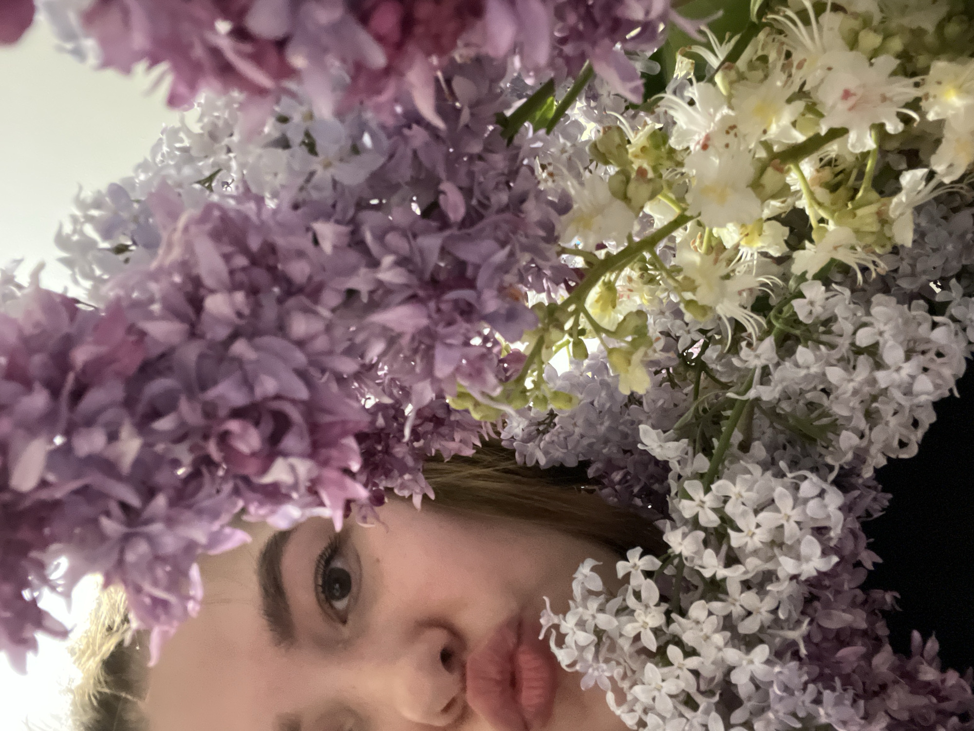 A person, presumably female based on facial features, is positioned beneath an arrangement of flowers, primarily lilacs in various shades of purple and white, along with some white horse chestnut blossoms. The person's face is visible from the eye level down, revealing an eye, nose, and pursed lips. The background behind the flowers appears to be a combination of light wall and dark area. This image does not provide enough information to accurately locate the scene in Novokuybyshevsk, Russia.