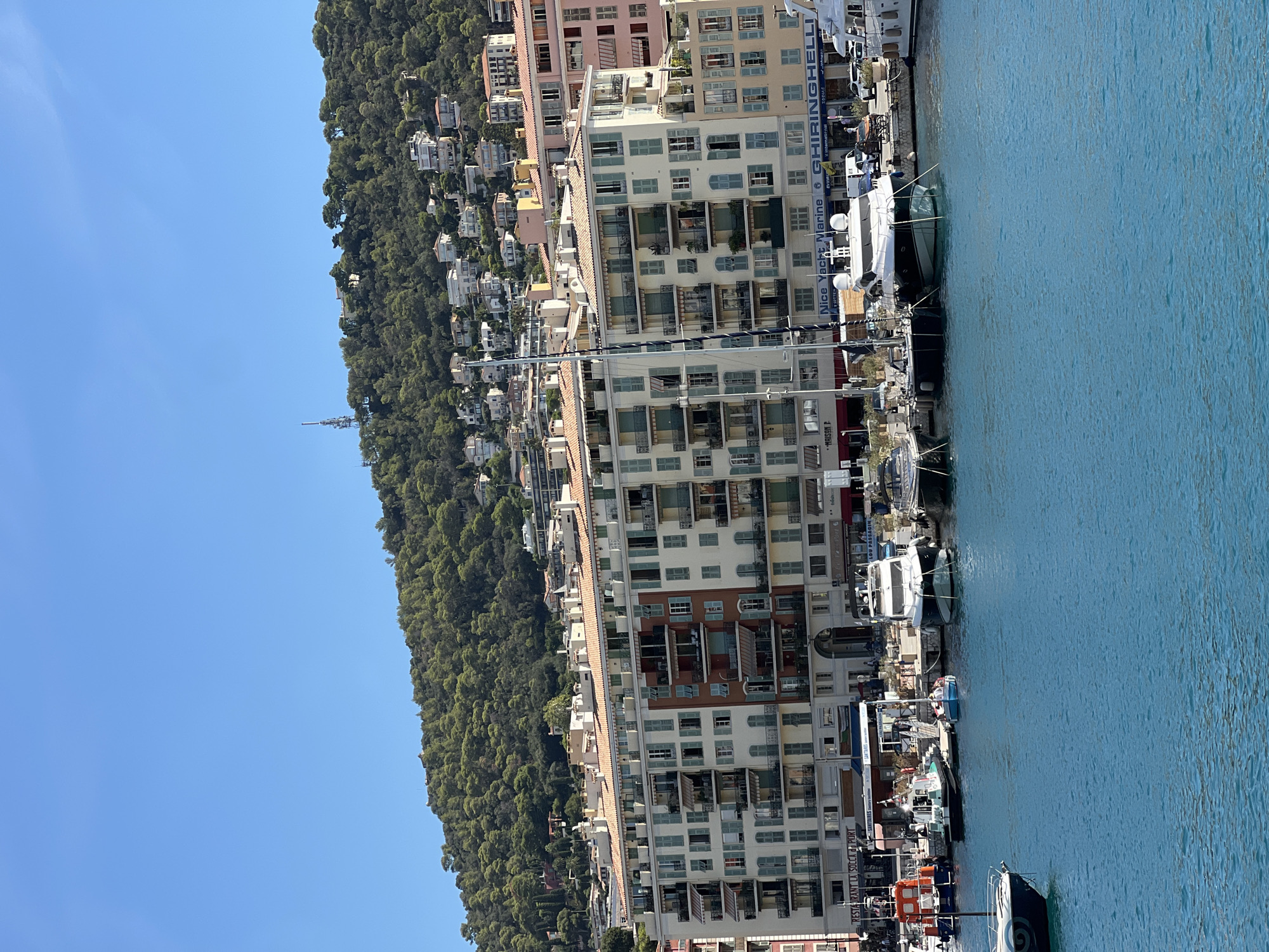 The image depicts a view of the port of Nice, France, on a clear, sunny day. In the foreground, tranquil blue water is visible, with several boats docked along the quay. Among them are a large white yacht, a smaller dark-hulled sailboat, and several other smaller vessels. The boats are moored and appear to be at rest.
Behind the boats, a long, multi-story building dominates the scene. The building is cream-colored with terracotta-tiled roofs and features numerous balconies with light-colored shutters. Some of the balconies have awnings extended. There are various businesses and restaurants located on the ground floor of this building, with visible signage such as "RESTAURANT LA SOCCA DU PORT," "Nice Yacht Marine," and "GHIRINGHELLI."
In the background, a densely wooded hill rises steeply behind the port buildings. The hillside is covered in a thick green forest. Scattered amongst the trees, and further up the slope, are numerous white and light-colored houses, indicative of a residential area on the elevated terrain. A telecommunications tower is visible near the crest of the hill. The sky above is a clear, bright blue, with no clouds present. The overall impression is one of a vibrant port city nestled against a natural, green landscape.