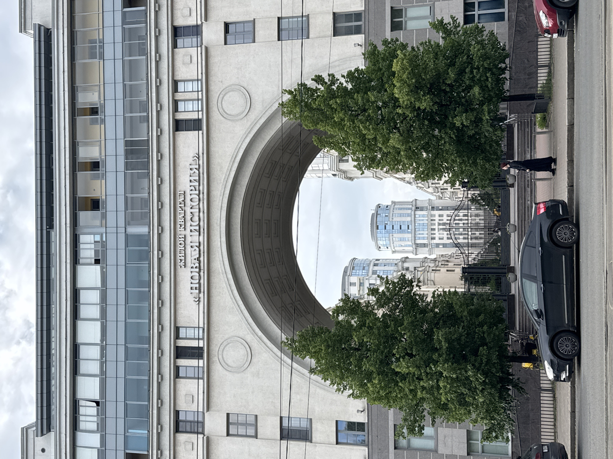 A low-angle, vertical shot captures a section of a modern building in Saint Petersburg, Russia. The building features a large, arched entryway with a curved, textured ceiling. Above the arch, an inscription in Russian reads "ЖИЛОЙ КВАPТАЛ "НОВАЯ ИСТОРИЯ"" which translates to "RESIDENTIAL BLOCK "NEW HISTORY"". To the left of the arch, a glass-covered facade of the building shows multiple floors with windows reflecting the overcast sky. To the right of the arch, lush green trees partially obscure a view of other buildings, including modern high-rises with glass facades. A black car is parked on the street next to a sidewalk where a person in a long black dress stands. The lighting suggests an overcast day.