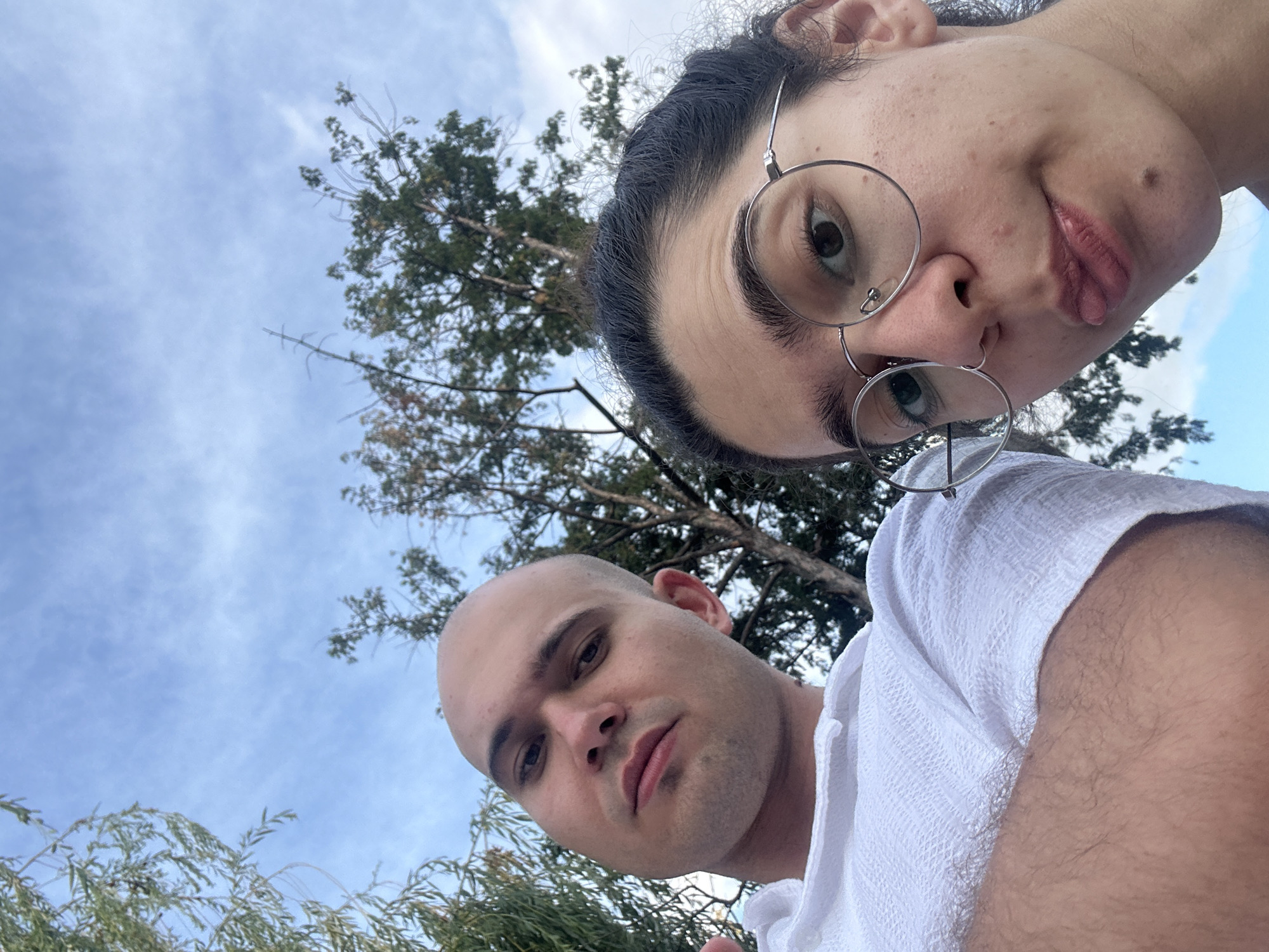 This is a close-up, low-angle selfie of two people outdoors, with a focus on their faces. The person on the left is a bald male wearing a white textured shirt, with his arm crossed. The person on the right is a female with dark hair pulled back, wearing round glasses and making a playful face, sticking her tongue out slightly. They are positioned in front of a background of trees and a cloudy sky. There is no discernible city, country, or specific location evident from the image itself, and therefore no information can be provided about Sofia, Bulgaria.