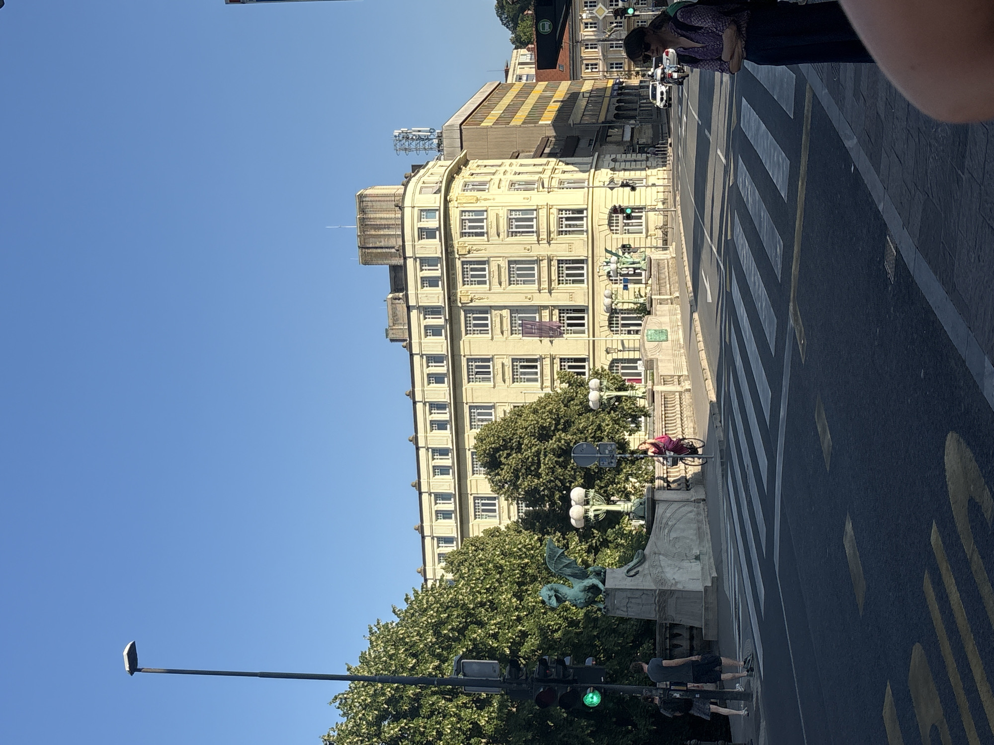 The image shows a street scene in a city. In the foreground, there is a pedestrian crossing. To the right, a woman wearing sunglasses and a patterned shirt stands with her arms crossed, facing away from the camera. In the middle ground, there is a large, pale yellow building with many windows. To the left of the building, a street light with a green signal is visible. A dragon statue is also visible on a bridge-like structure. A person is cycling on the bridge, wearing a pink outfit. Several trees with green foliage are present. In the background, other buildings and vehicles are visible. The sky is clear and blue.