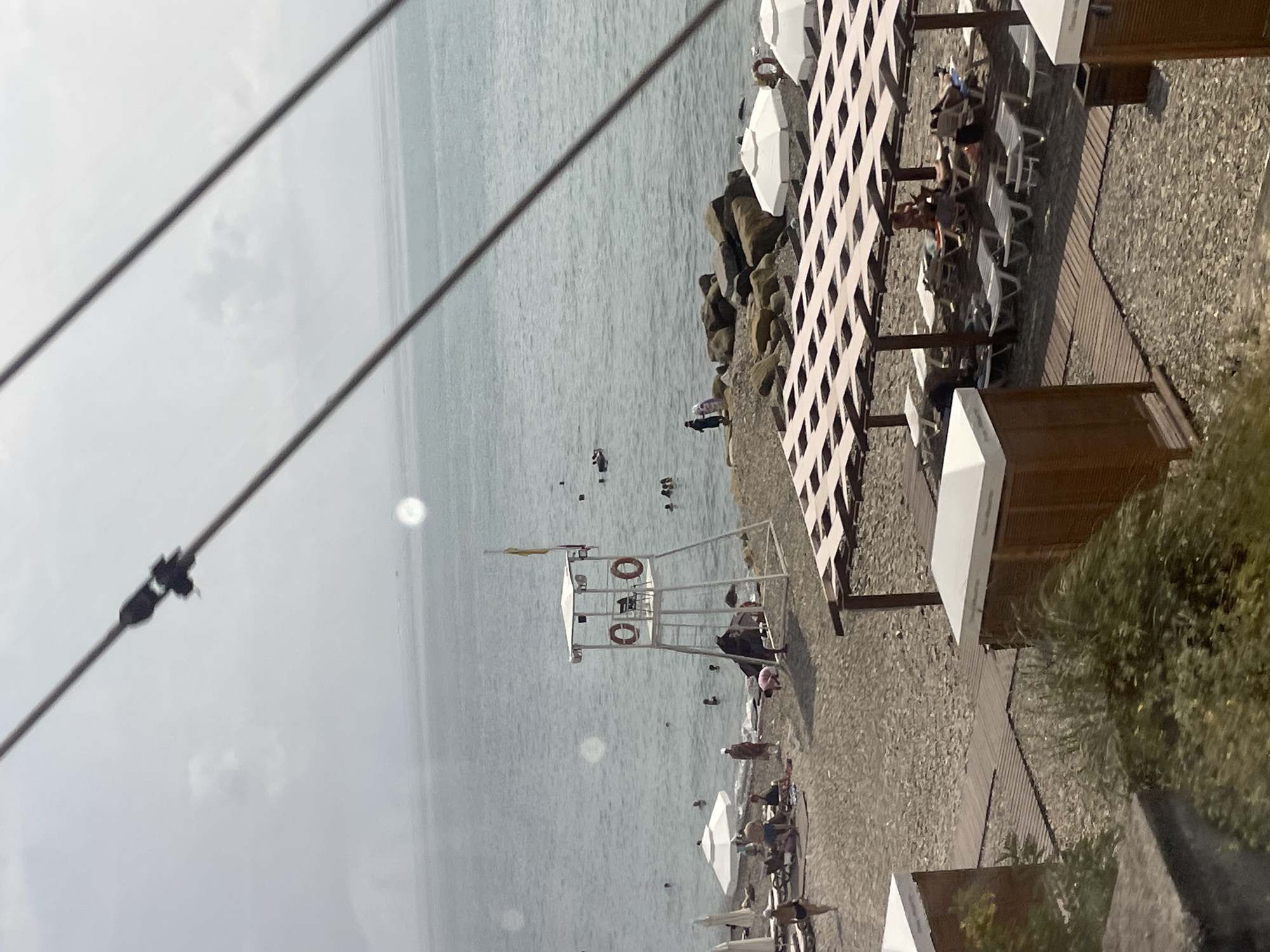 The image depicts a cloudy day at a beach in Sochi, Russia. The scene is a section of a beach with scattered individuals engaged in various activities. In the foreground, two wooden structures with white roofs and closed wooden shutters are visible, alongside a wooden walkway. Further back, a beach umbrella provides shade over several lounge chairs occupied by people relaxing. The beach itself is a mix of sand and pebbles.
In the middle ground, a white lifeguard tower stands prominently near the water's edge. It features a ladder, a small platform, and two life preservers. Several people are scattered on the beach and in the shallow water. Some are sunbathing on lounge chairs, while others are wading or swimming. The sea is calm with gentle waves.
In the background, the vast expanse of the sea meets the hazy sky. A rocky outcropping is visible to the right of the lifeguard tower. The overall atmosphere is one of a typical day at a seaside resort, with a slightly overcast sky suggesting cooler temperatures or a break from intense sun.
