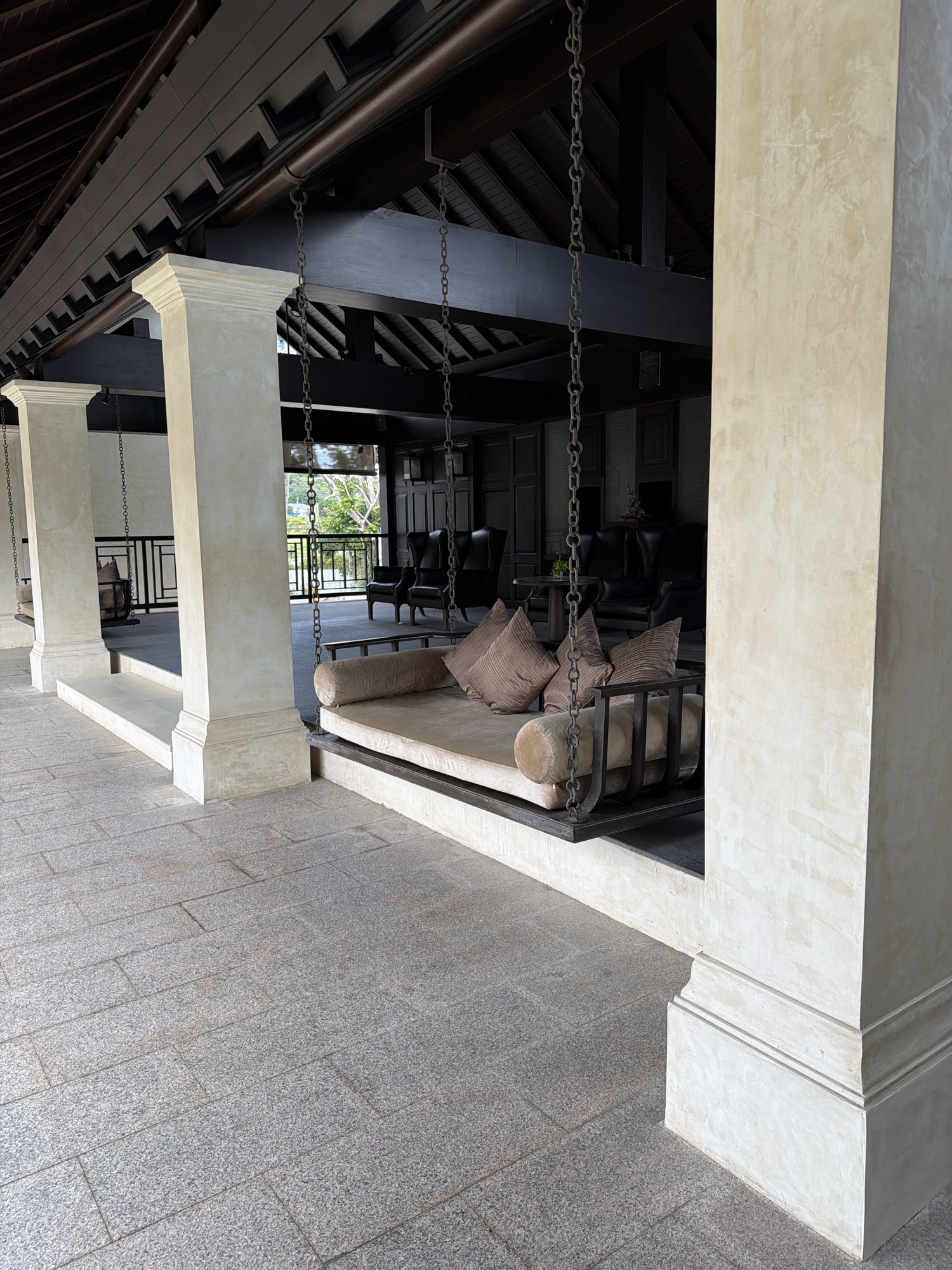 The image shows a covered outdoor seating area with a large swing bed and several chairs. The swing bed is made of a light-colored fabric with brown cushions and is suspended by chains from the dark wooden ceiling structure. To the left, two more swing beds are visible, also suspended by chains. Behind the swing bed, a seating area is furnished with dark leather chairs and a small round table. The floor is paved with gray granite tiles. Large, light-colored rectangular columns support the roof structure. The scene appears to be an outdoor lounge area, possibly at a resort, in a tropical setting given the lush greenery visible through a railing in the background. The overall aesthetic is one of relaxed elegance, with dark wood and light plaster creating a sophisticated ambiance. This location is identified as Ko Samui District, Thailand, suggesting a luxurious tropical resort or hotel setting. There are no people visible in the image.