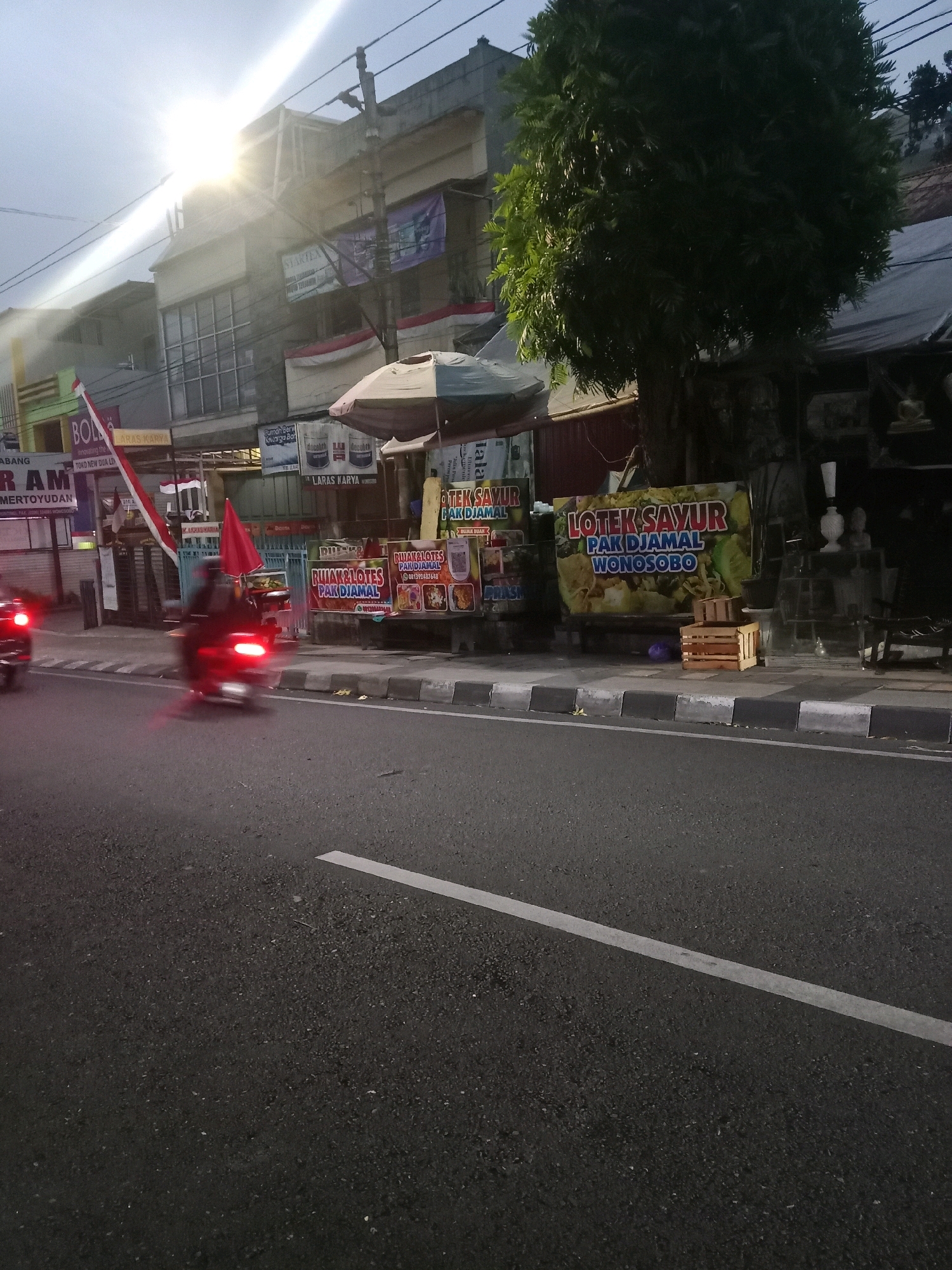 The image depicts a street scene in Wonosobo, Indonesia, likely during the early morning or late afternoon due to the low light and sun glare. A road with a white line marking the lane is visible in the foreground. On the right side of the road, a sidewalk runs alongside buildings. A large, leafy tree dominates the middle ground on the right, casting shadows. A series of food stalls are set up on the sidewalk, featuring various signs. One prominent sign reads "LOTEX SAYUR PAK DJAMAL WONOSABO," indicating a local food vendor. Another sign says "RUJAK LOTES PAK DJAMAL." Several wooden crates are stacked on the sidewalk near the stalls. On the left side of the street, a motorcycle with its taillights illuminated is captured in motion, suggesting traffic. Further back, buildings with multiple floors are visible, some with signs like "STAR TEX" and "LARAS KARYA." The overall atmosphere is that of a bustling urban environment with commercial activity.