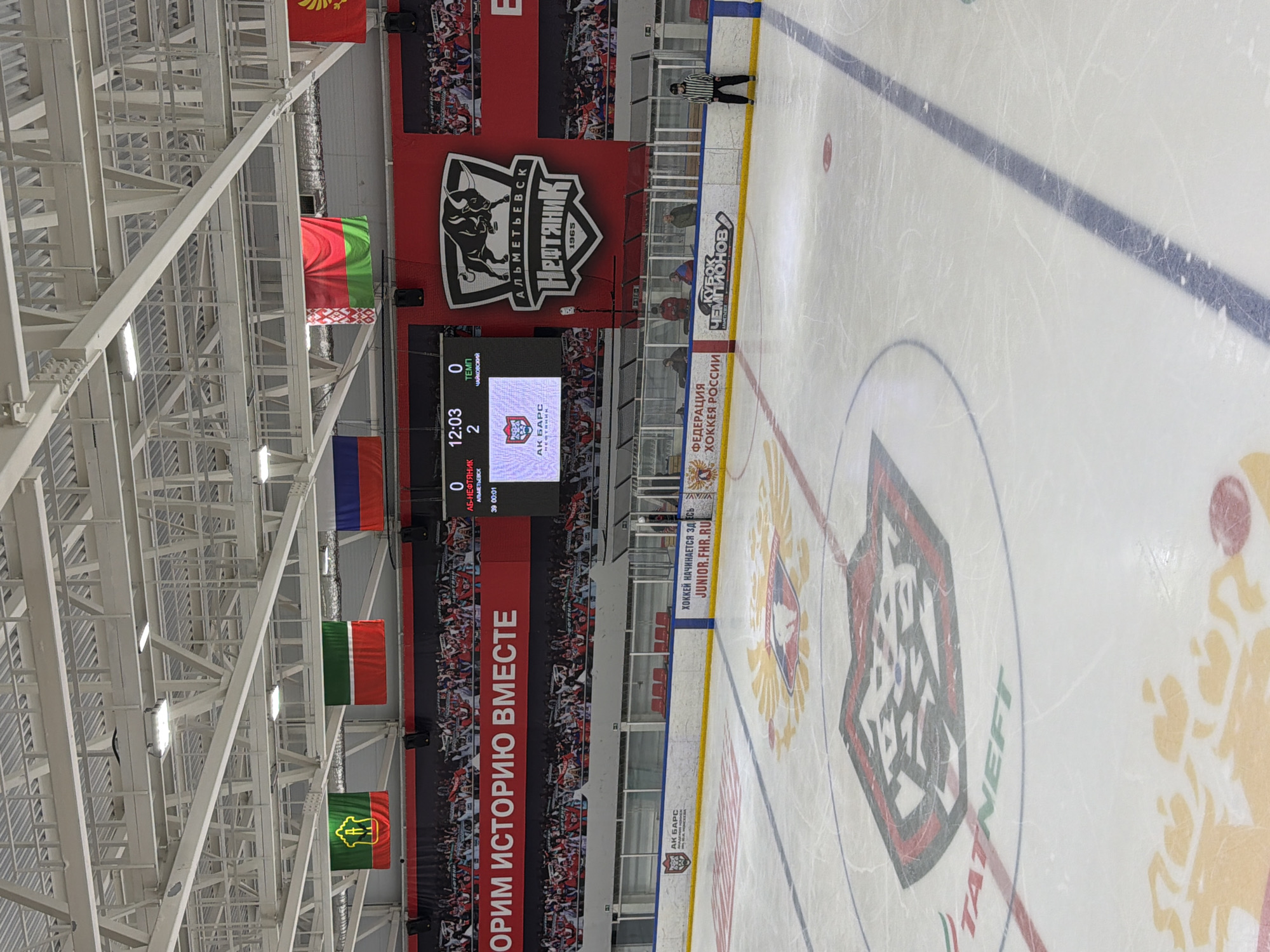 This image shows a view inside a hockey arena, potentially in Almetyevsk, Russia, based on the scoreboard displaying "АБ-НЕФТЯНИК АЛЬМЕТЬЕВСК" and the "TATNEFT" logo on the ice. The score is 0-2, with the time showing 12:03 in the first period. Banners hang in the background, including one with the team logo "Нефтяник Almetyevsk 1965." There is a referee in black and white stripes standing near the boards. The ice has logos for "Федерация хоккея России" (Russian Ice Hockey Federation) and "Кубок чемпионов". Flags of different countries are suspended from the ceiling. The words "ОРИМ ИСТОРИЮ ВМЕСТЕ" are on the banner.