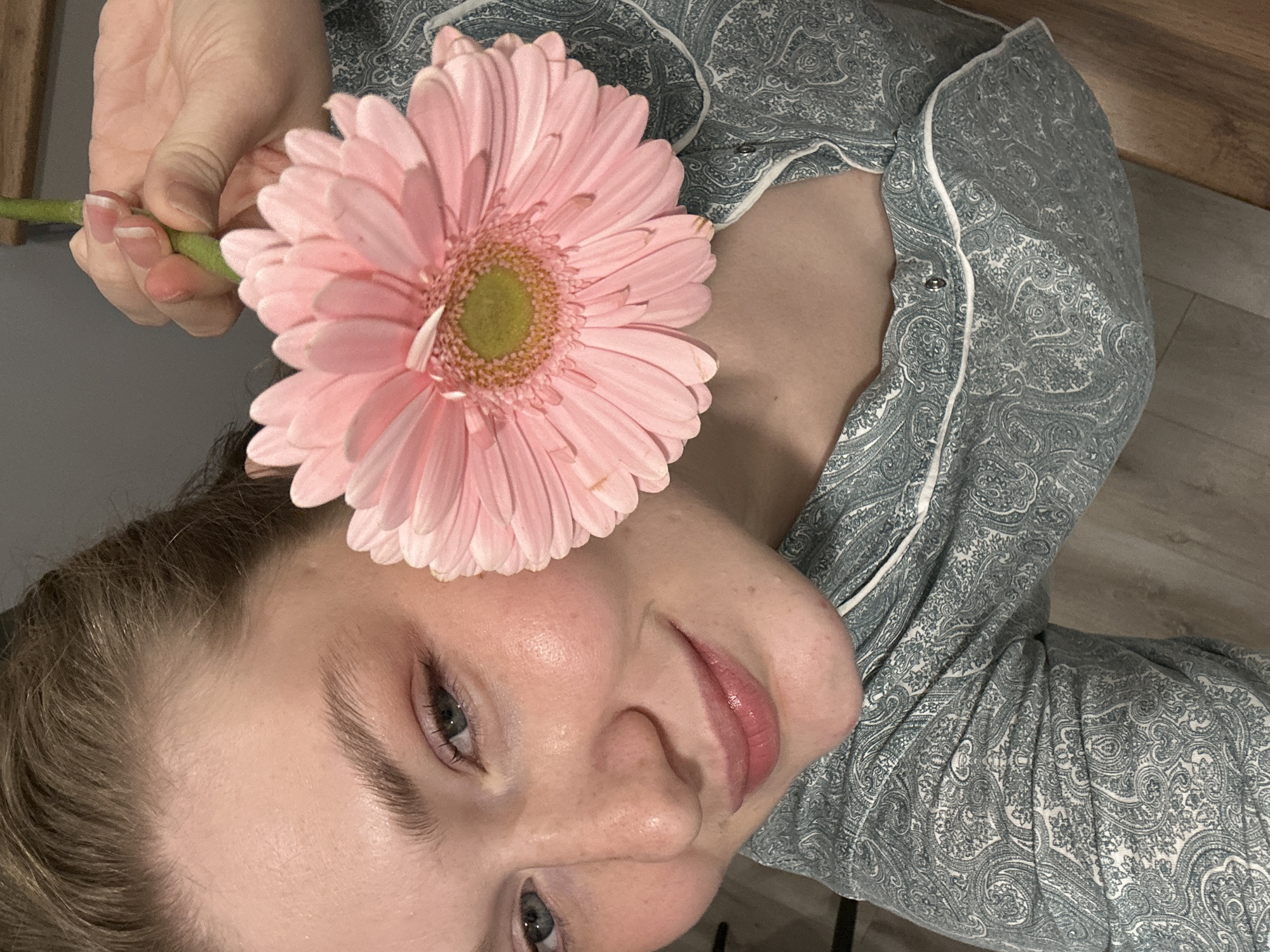 A woman in a blue and white paisley patterned top is holding a pink gerbera daisy flower in her hand. The flower is positioned near her forehead. The woman's face is angled towards the camera, and she is smiling slightly. Her eyes are light-colored. The background consists of a wooden surface and a light-colored floor. It's impossible to know with certainty where this photo was taken, but there is no evidence here to suggest the location is Kazan, Russia.