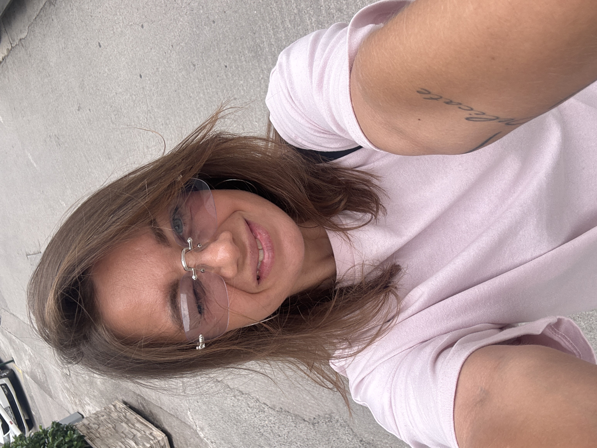 A portrait-oriented selfie features a smiling young woman with long, light brown hair, captured outdoors in bright daylight. She wears a light pink short-sleeved t-shirt and rimless, light-tinted sunglasses with silver bridge and temple details. Her expression is joyful, with a broad smile revealing her teeth, and she has a small, decorative nose piercing.
Her left upper arm is visible, displaying a tattoo that clearly reads "delicate" in a cursive script font. The background primarily consists of a light gray concrete or asphalt surface, suggesting an urban outdoor setting such as a street, sidewalk, or plaza. In the blurred lower left of the frame, elements resembling a white vehicle and green foliage from a planter are faintly visible. The overall mood is casual and happy, consistent with a leisurely moment in a location like Budva, Montenegro.
